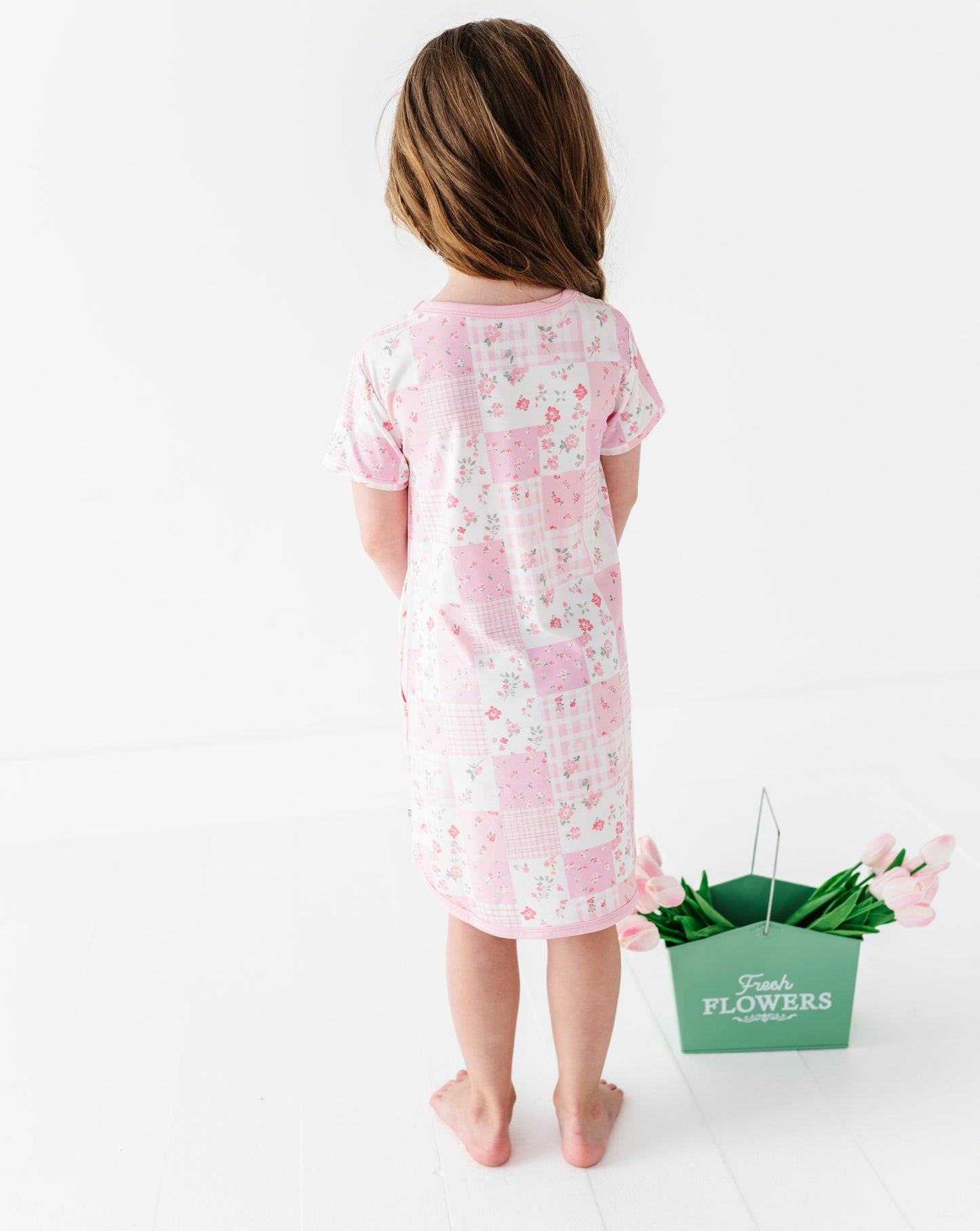 Child wearing a pink floral bamboo gown standing next to a green bucket of flowers on a white background