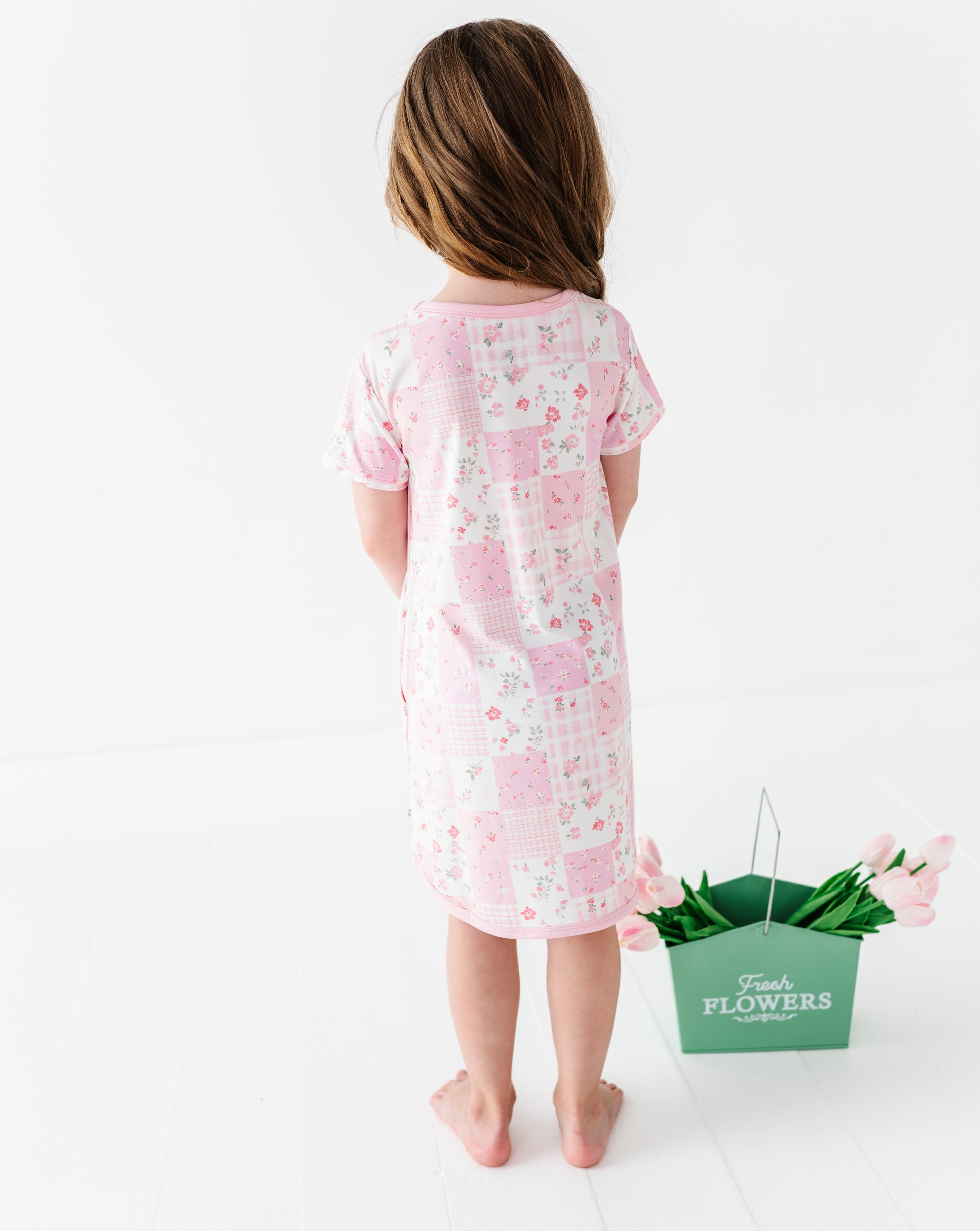 Child wearing a pink floral bamboo gown standing next to a green bucket of flowers on a white background