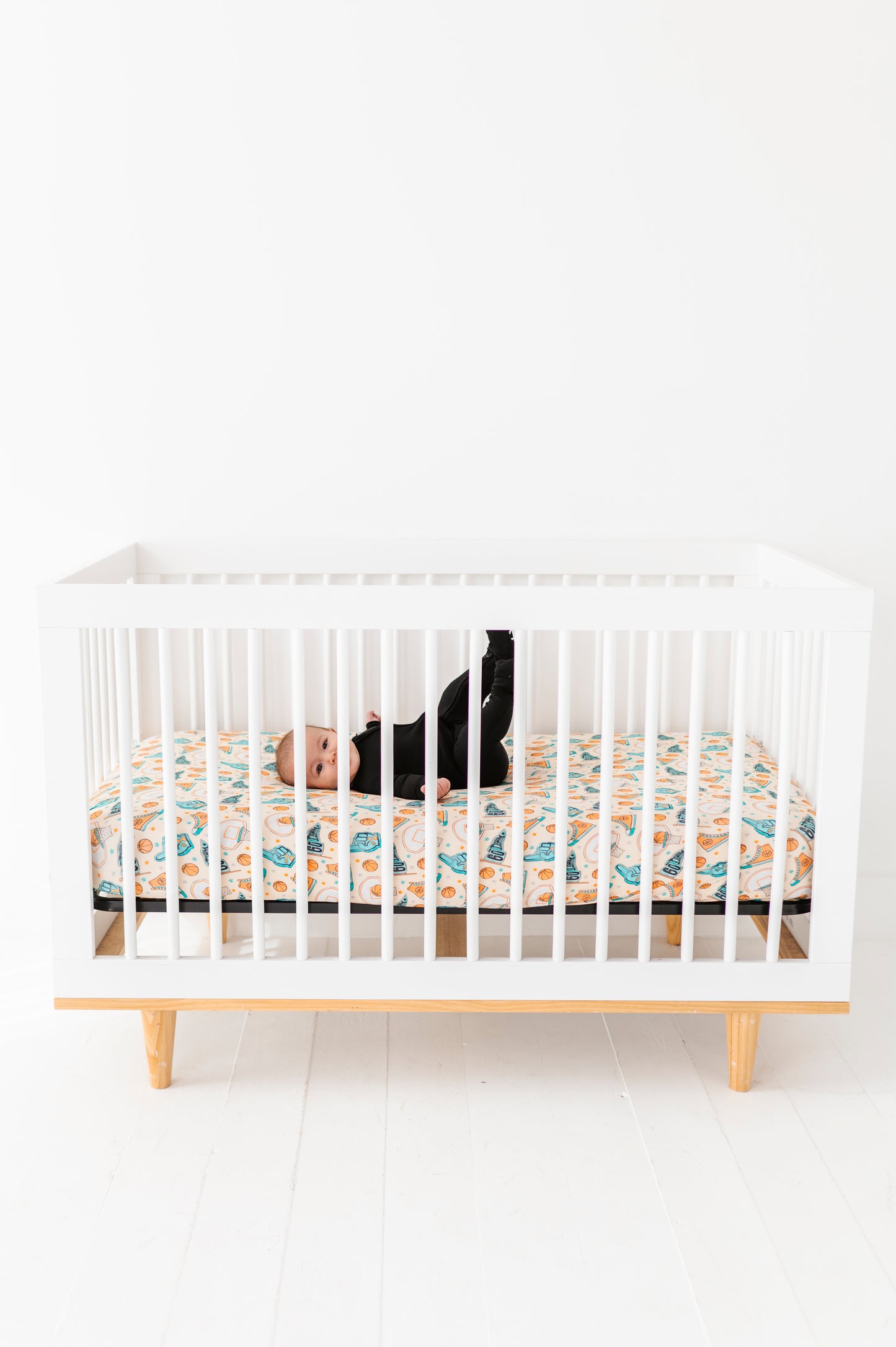 Baby laying in a white crib with wooden legs featuring a basketball themed bamboo crib sheet