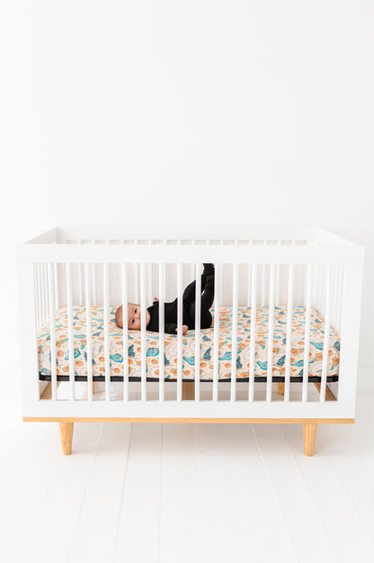 Baby laying in a white crib with wooden legs featuring a basketball themed bamboo crib sheet