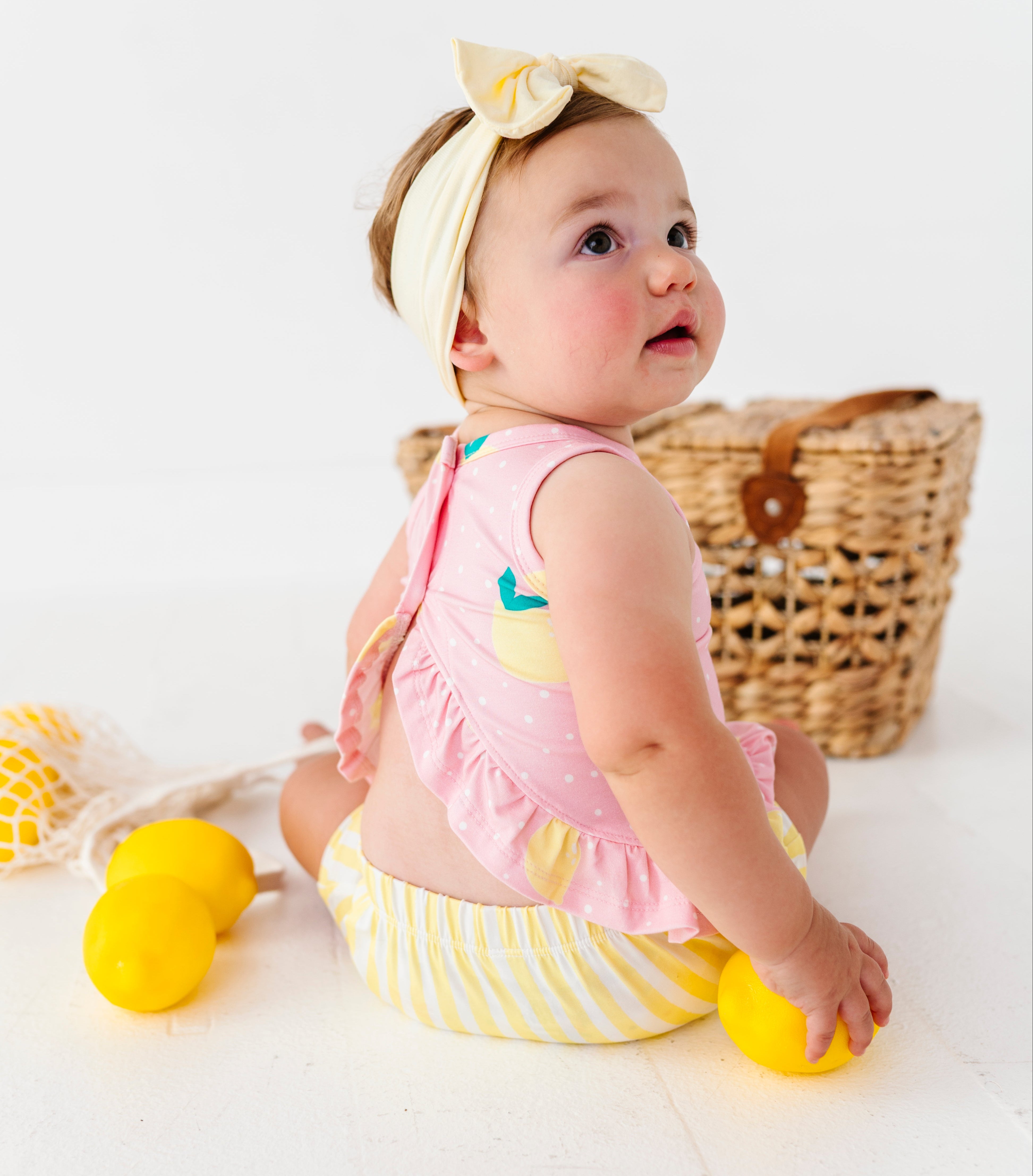 Baby in pink top and yellow striped shorts sitting on a white surface with lemons and a basket.
