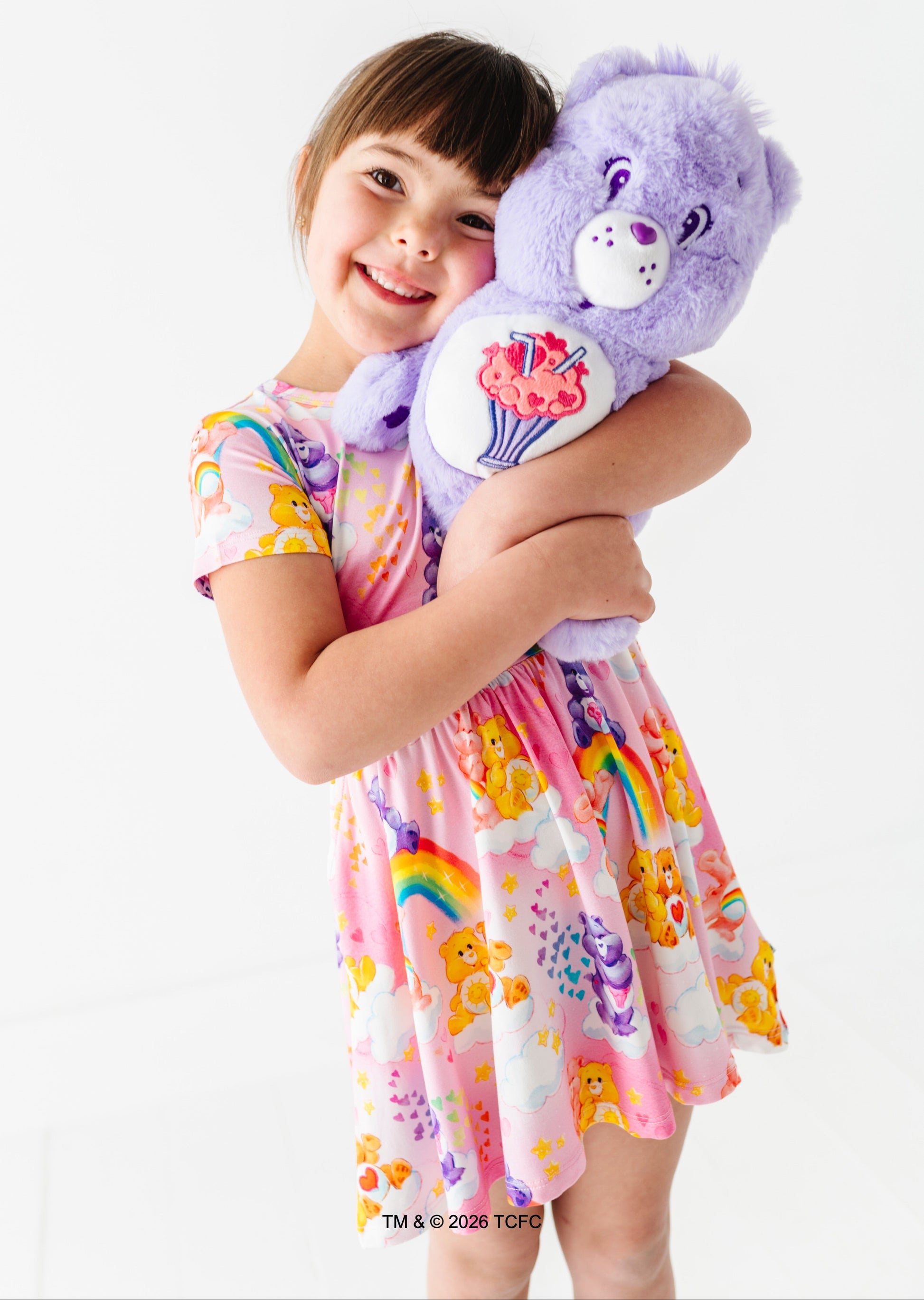 Child in a colorful bamboo dress holding a purple Care Bears™ plush on a white background