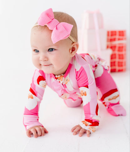A baby crawling on a white surface wearing a pink Santa-themed footed bamboo onesie and a matching pink bow headband, with wrapped gifts blurred in the background.