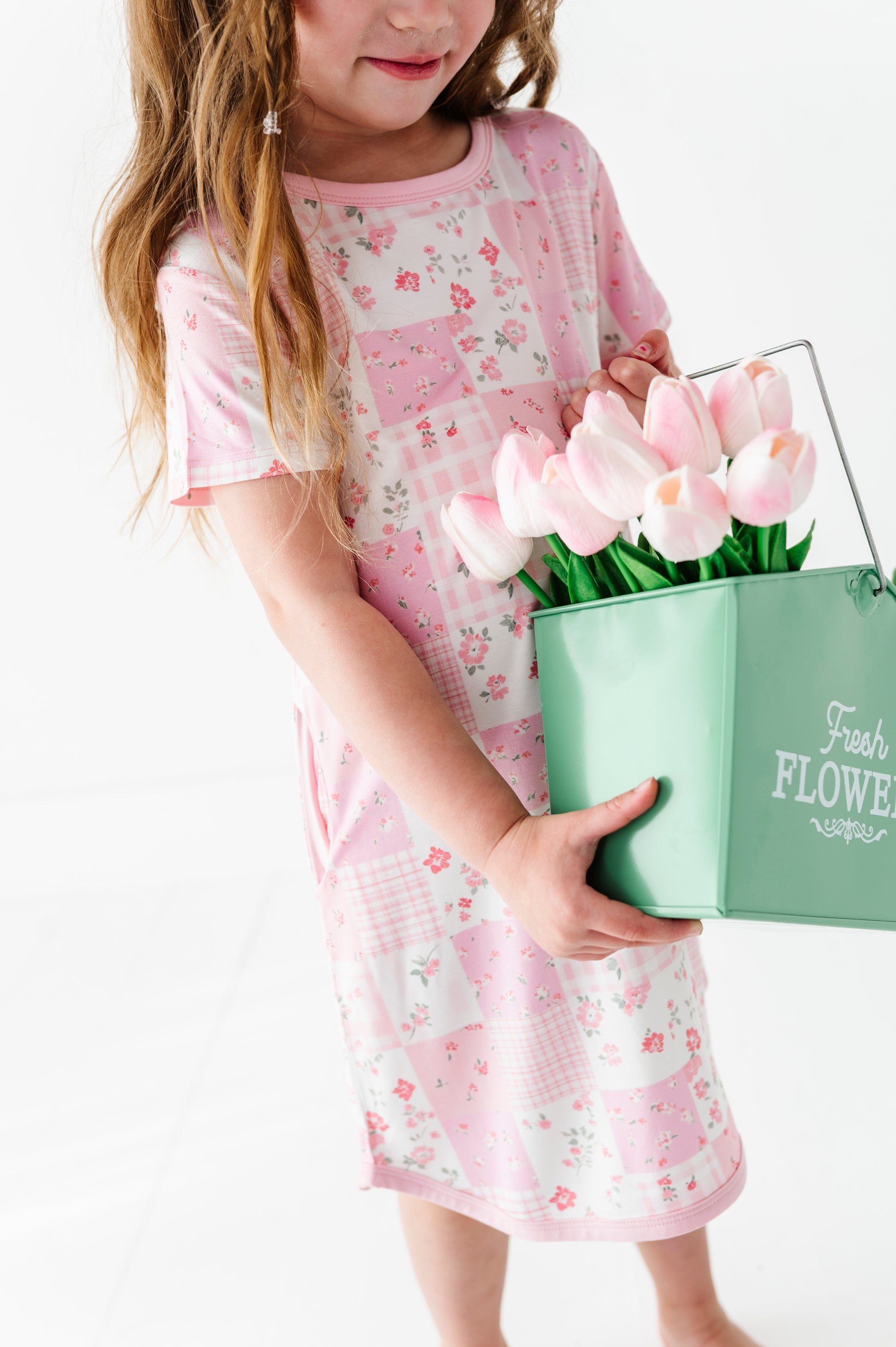 Child in a pink floral gown holding a green bucket of tulips on a white background