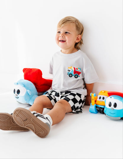 A toddler sitting on a white floor against a white wall, wearing a white graphic t-shirt with a colorful truck design, black and white checkered shorts, and brown sneakers. Colorful plush toys are placed around the child.