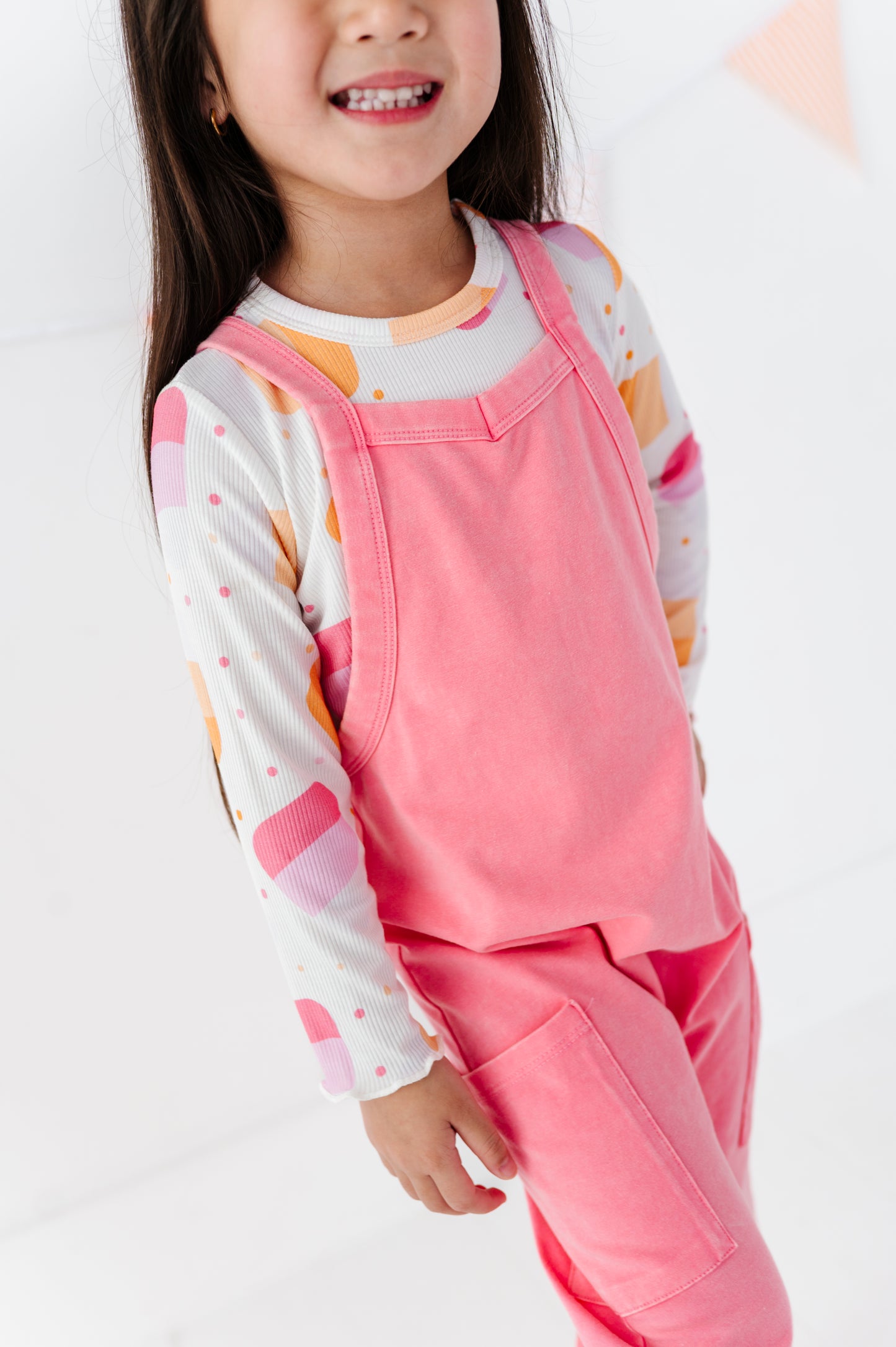 A young girl wearing a pink overall jumpsuit over a white long-sleeve shirt with pink, orange, and yellow geometric patterns, shown in a bright studio setting.