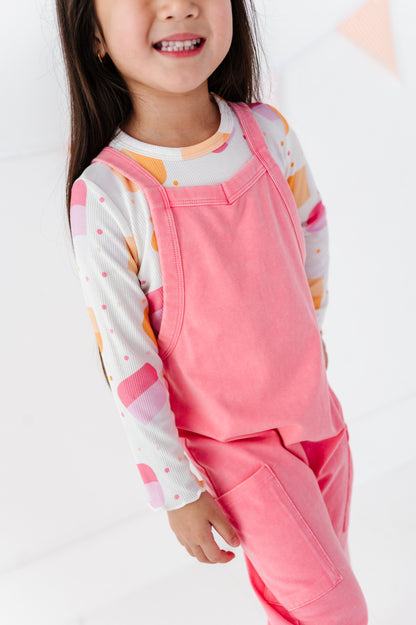 A young girl wearing a pink overall jumpsuit over a white long-sleeve shirt with pink, orange, and yellow geometric patterns, shown in a bright studio setting.