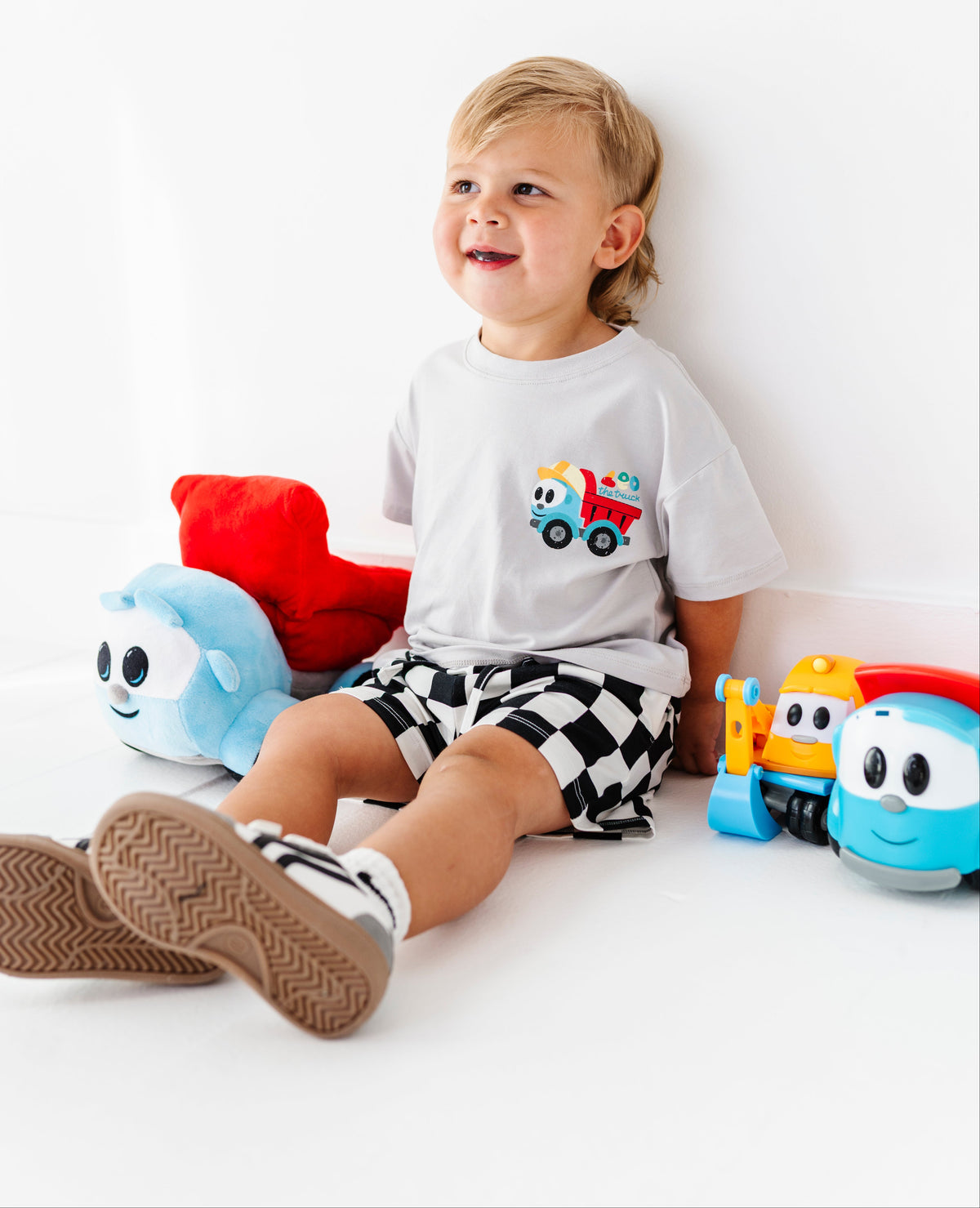 A toddler sitting on a white floor against a white wall, wearing a white graphic t-shirt with a colorful truck design, black and white checkered shorts, and brown sneakers. Colorful plush toys are placed around the child.