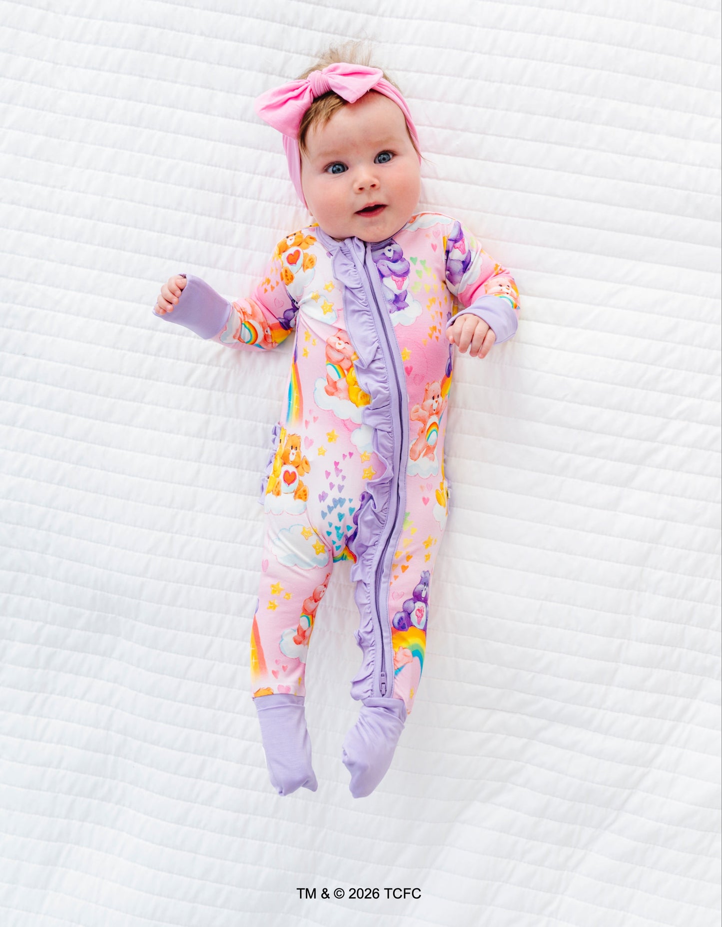 Baby in a colorful onesie with a pink bow on a white textured surface