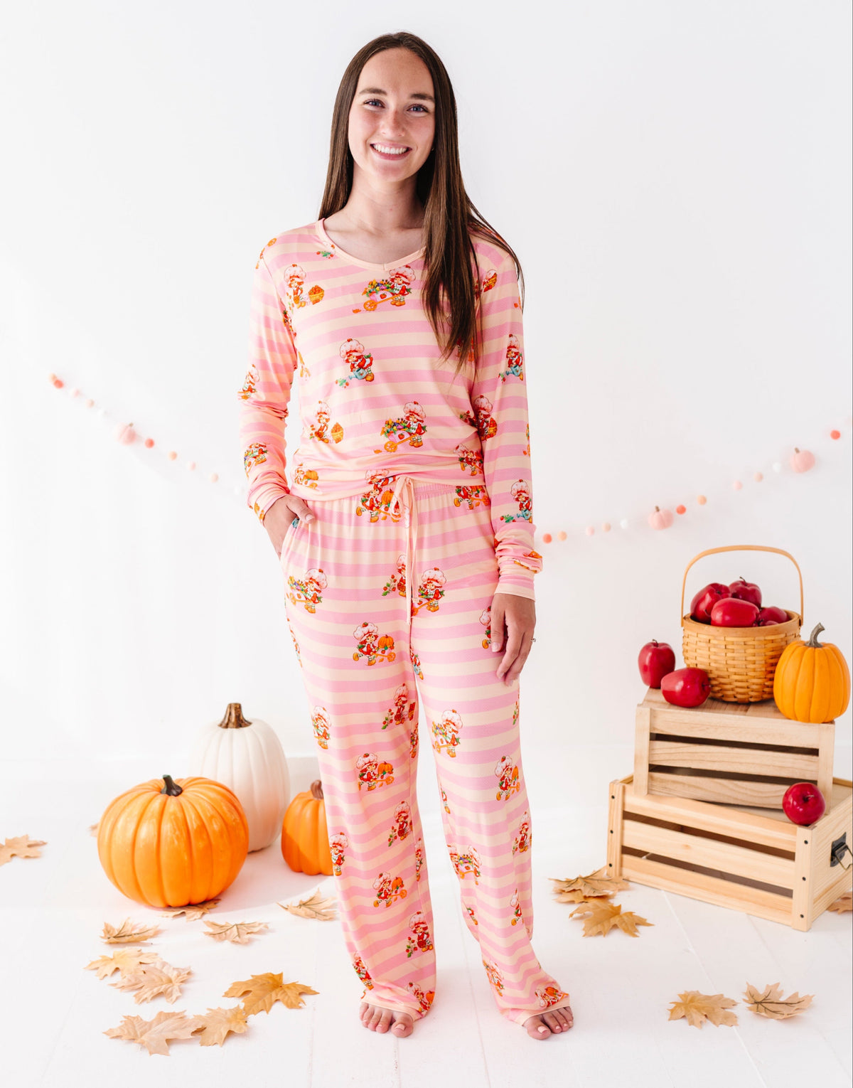 A woman stands barefoot in a studio wearing a pink pajama set with orange and yellow autumn motifs. The set includes a long-sleeve top and matching pants.