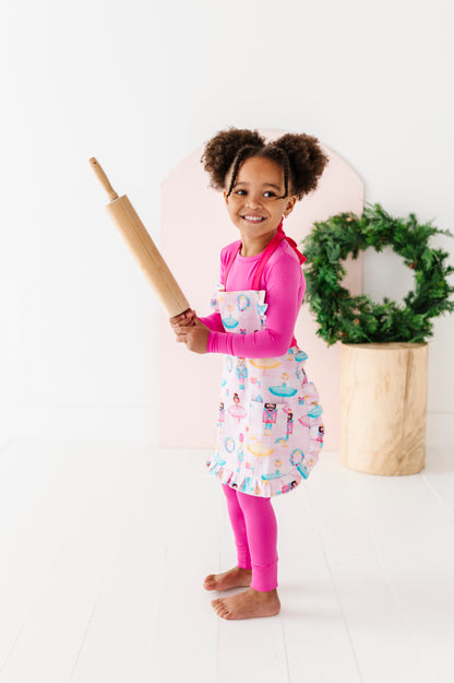 Child wearing a light pink Christmas apron holding a rolling pin with a wreath in the background.