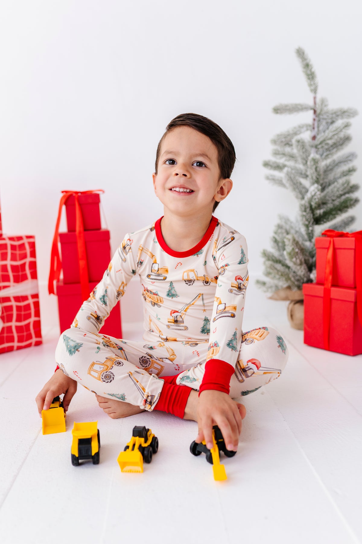 A little boy sitting on a white floor playing with construction trucks while wearing construction-themed bamboo pajamas 