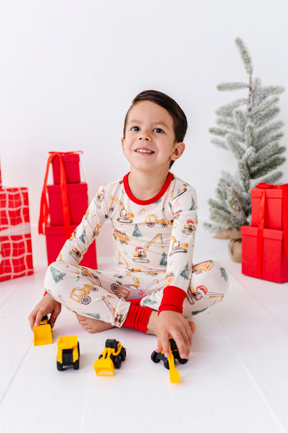 A little boy sitting on a white floor playing with construction trucks while wearing construction-themed bamboo pajamas 