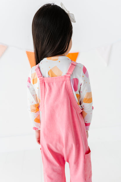 A young child viewed from behind wearing a pink overall-style jumpsuit over a white long-sleeve shirt with orange and pink abstract shapes, against a bright white indoor background.