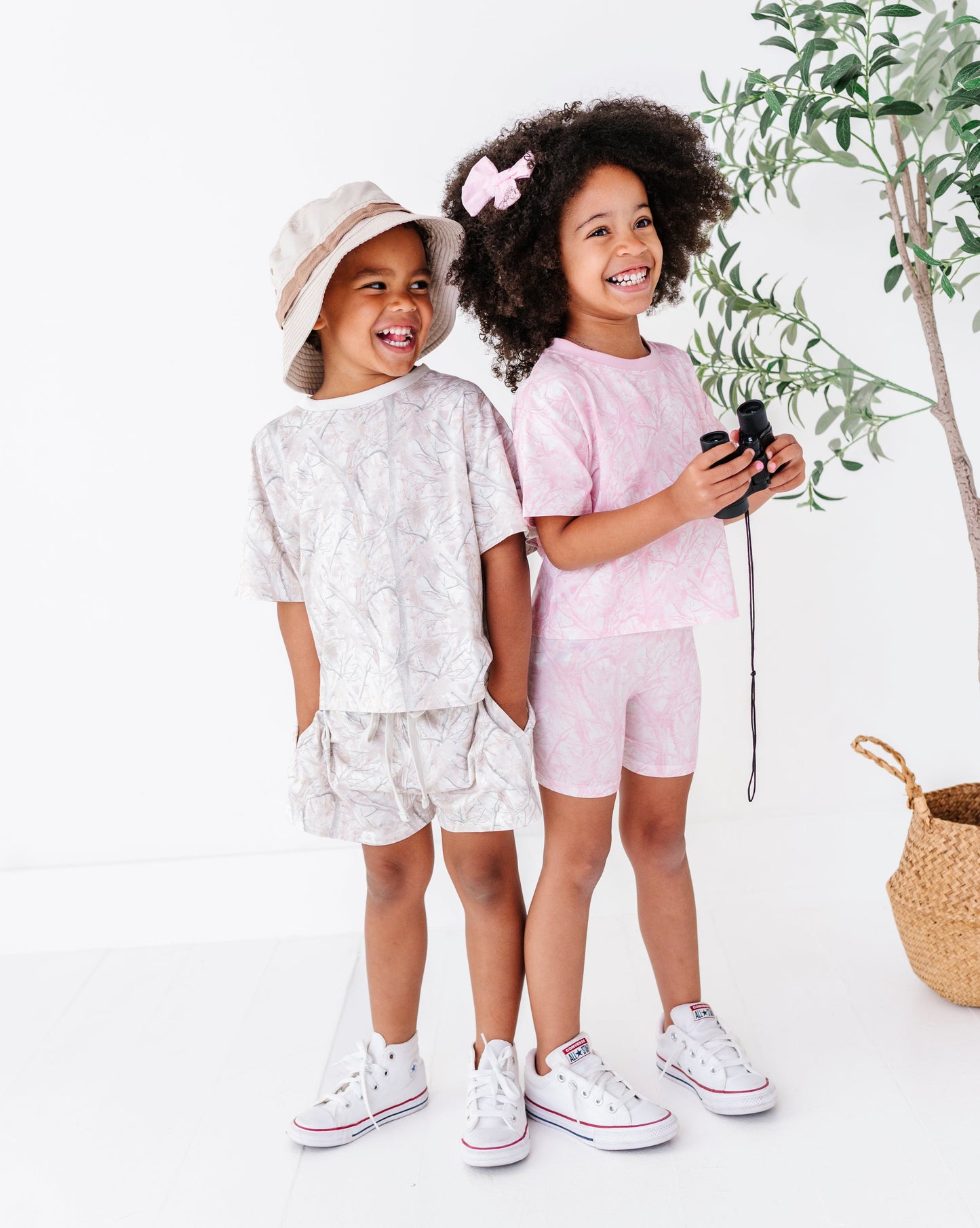 Two children in matching outfits standing next to a plant on a white background