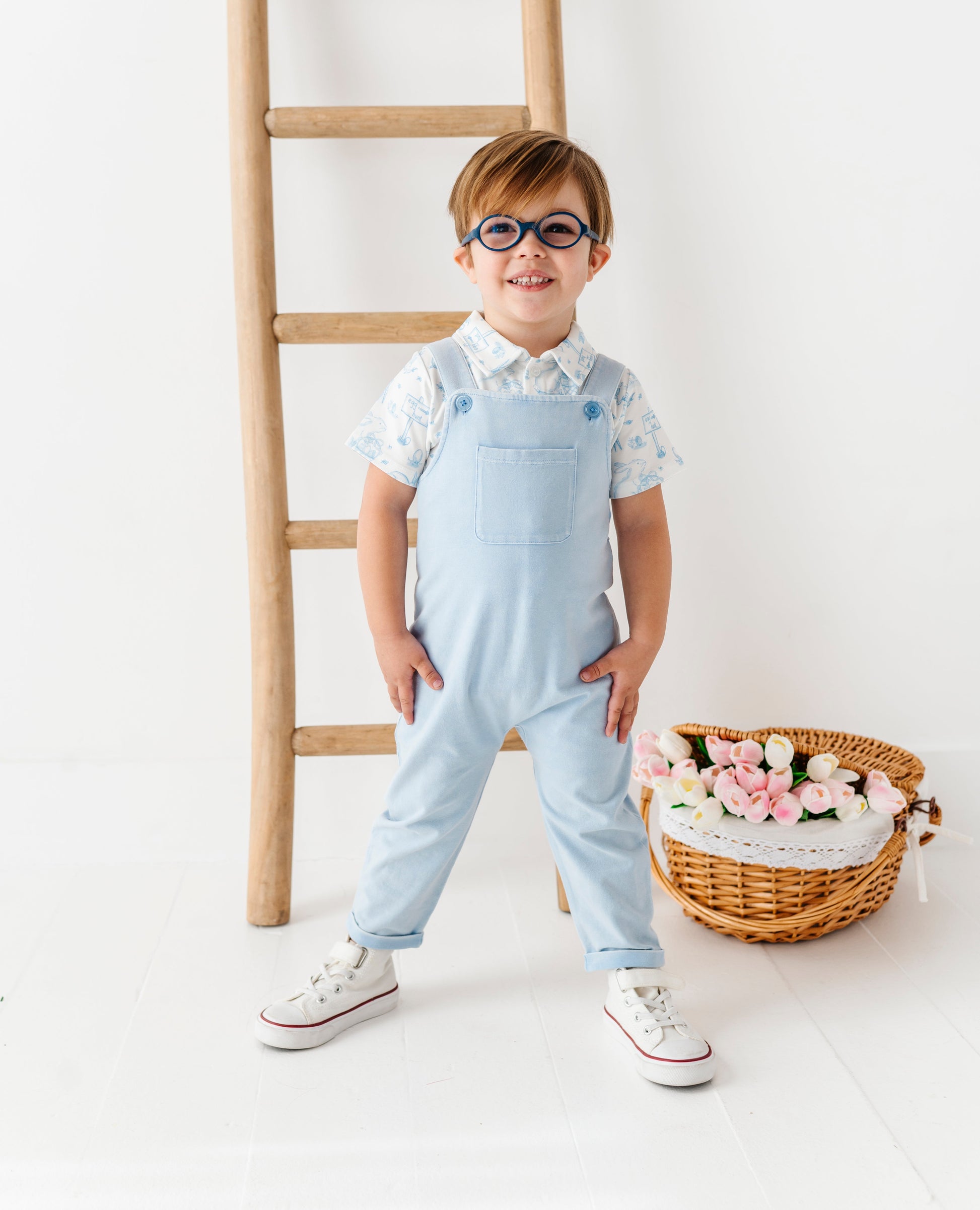 Child wearing light blue overalls standing next to a wooden ladder and flower basket on a white background