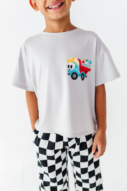 A smiling child wearing a white short-sleeve T-shirt with a colorful cartoon dump truck graphic on the chest, paired with black-and-white checkered pants, against a plain white background.