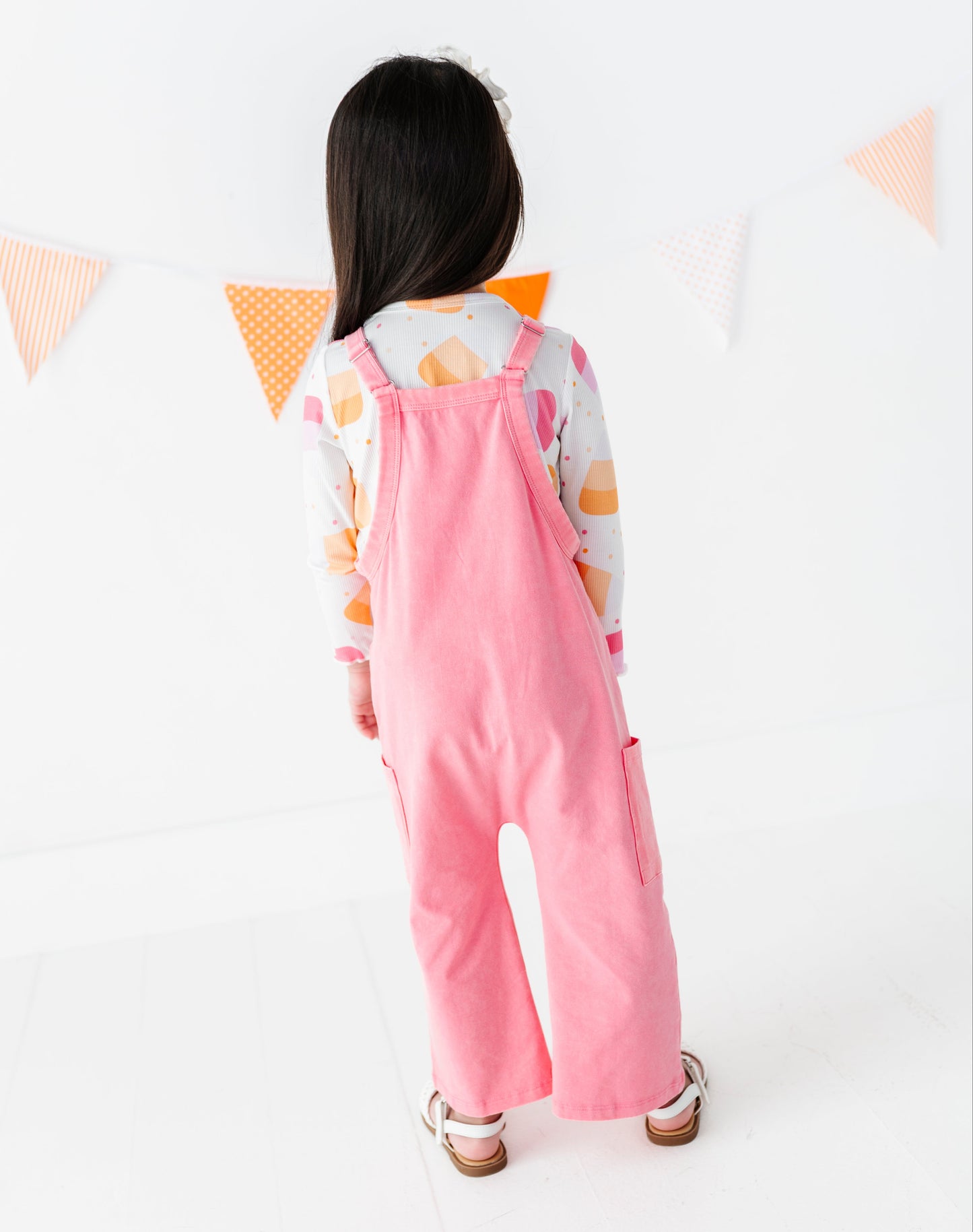 A young child viewed from behind wearing pink corduroy overall-style pants over a white long-sleeve shirt with a pastel fruit print, standing against a white wall with orange and white bunting.