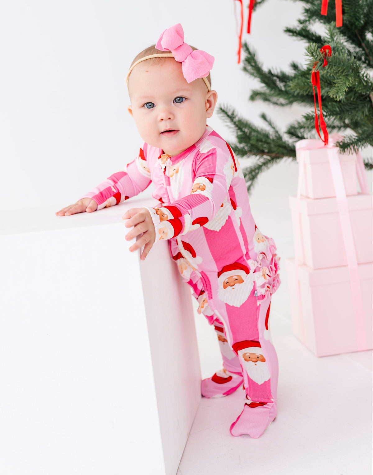 A baby wearing a pink Santa Claus print footed bamboo pajama onesie and a matching pink bow, leaning on a white box in a studio setting with a Christmas tree and wrapped gifts in the background.