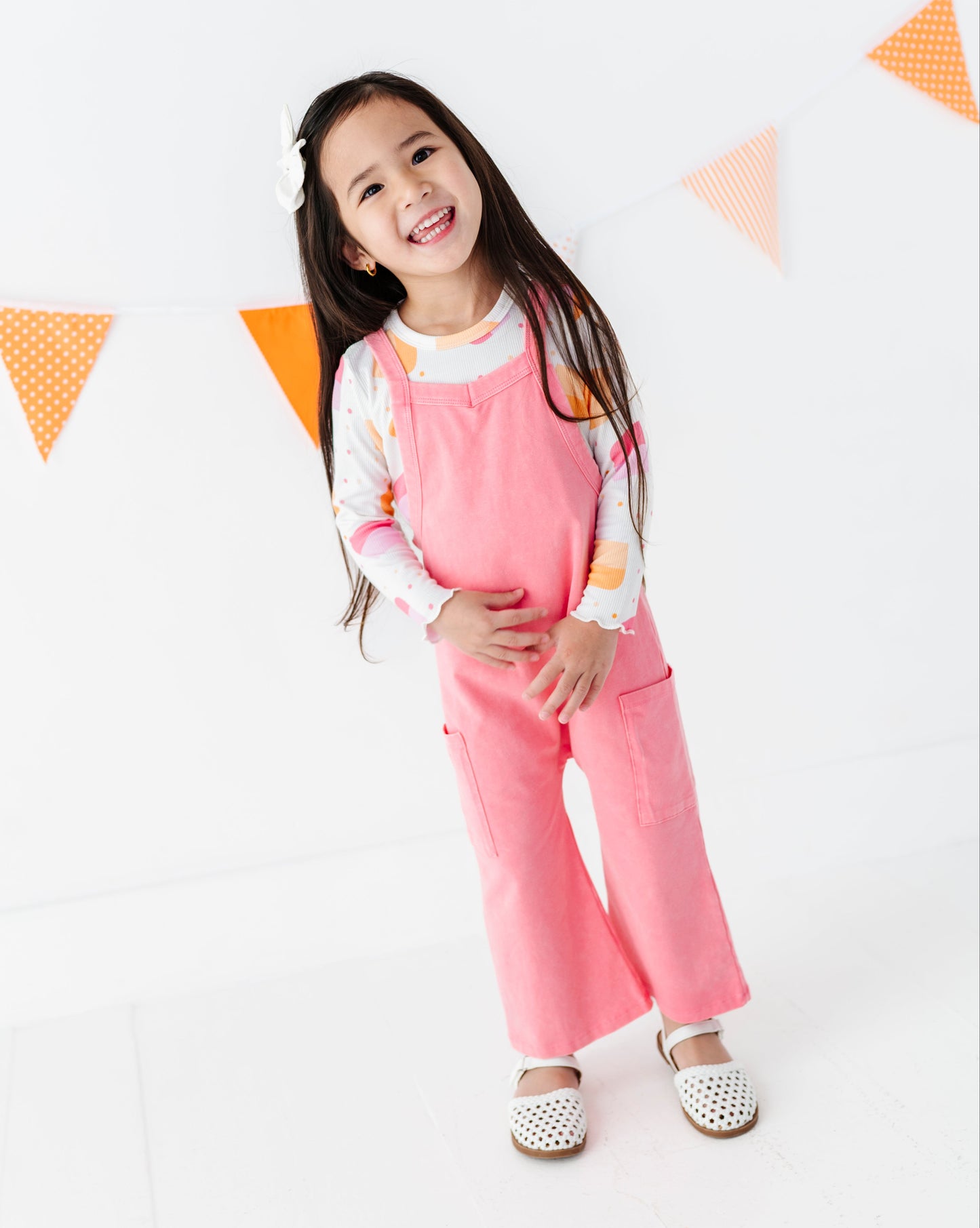 A young girl smiling and posing in pink overall pants and a white long-sleeve shirt with orange accents, standing against a white wall with orange bunting.