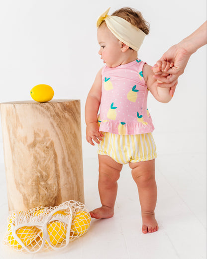Baby in pink sleeveless top and yellow striped shorts holding a hand, with a wooden block and lemon in the background.