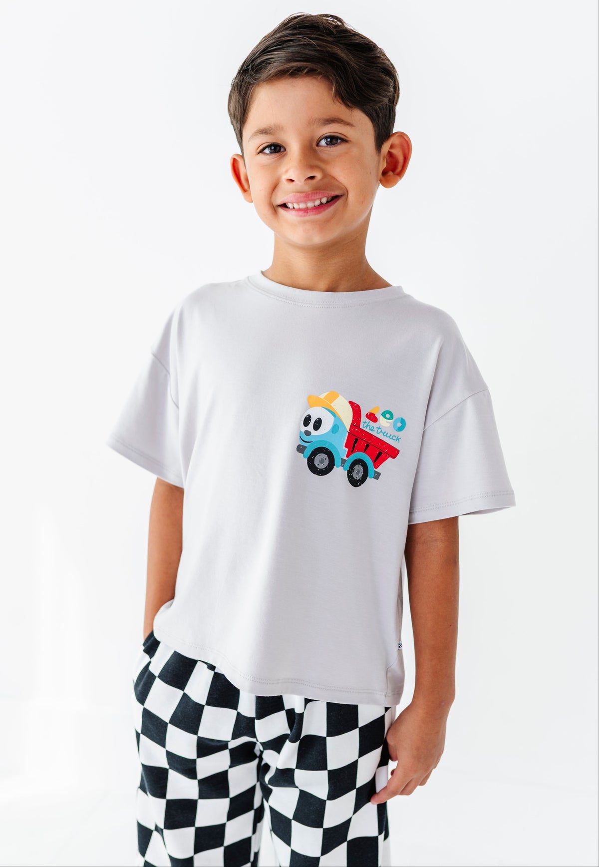 A smiling young boy wearing a white short-sleeve t-shirt with a colorful cartoon dump truck graphic on the chest, paired with black and white checkered pants, against a plain white background.