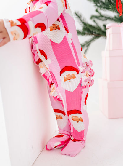 A baby wearing a pink footed one-piece bamboo pajama with a Santa Claus print, leaning against a white wall in a holiday-themed setting.