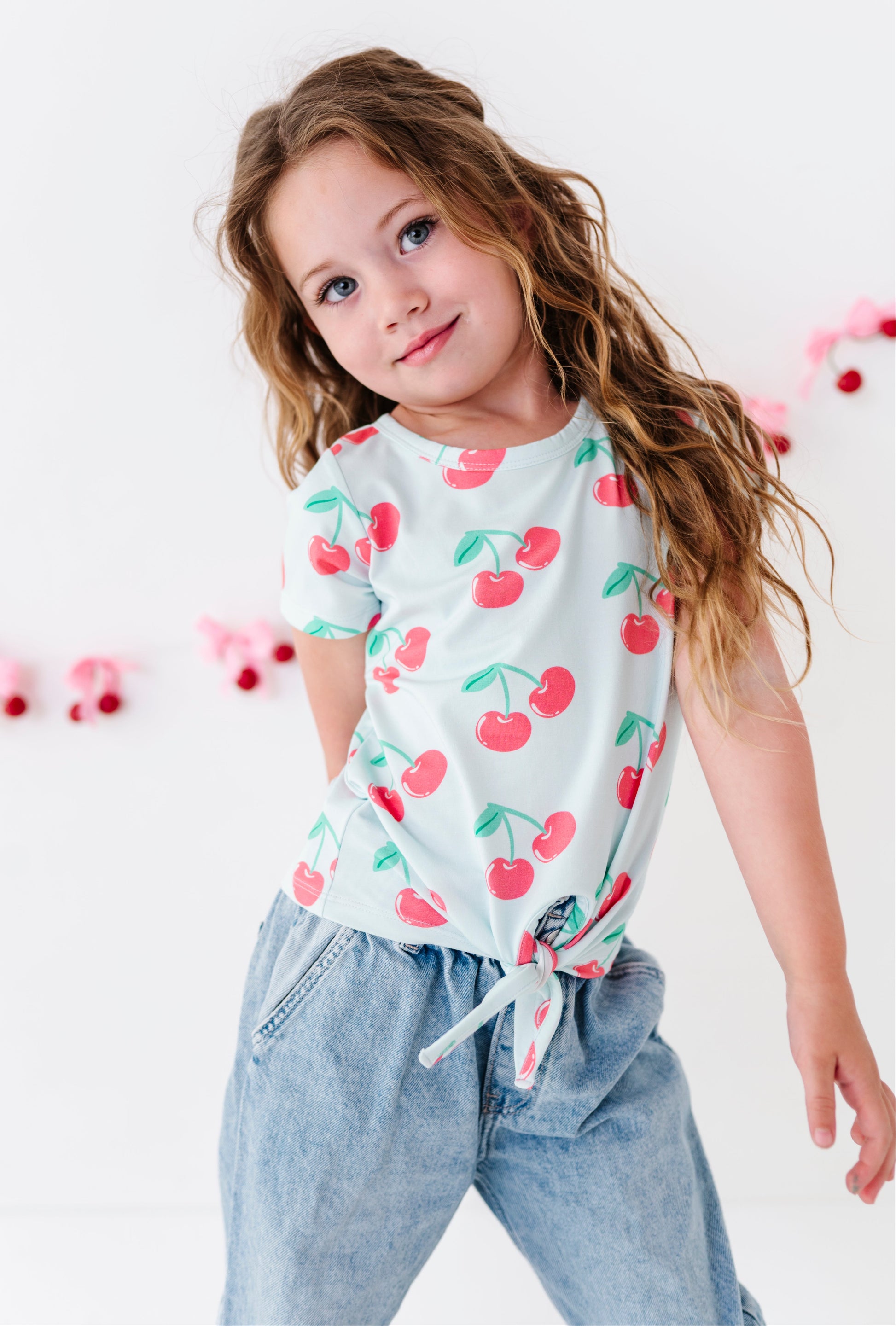 A young girl with long wavy brown hair wearing a white short-sleeve t-shirt with a red cherry print and light blue jeans, posing against a white wall with pink decorative garlands.