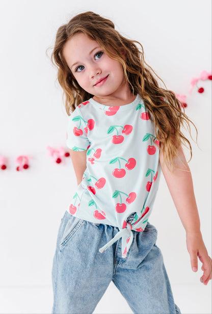 A young girl with long wavy brown hair wearing a white short-sleeve t-shirt with a red cherry print and light blue jeans, posing against a white wall with pink decorative garlands.
