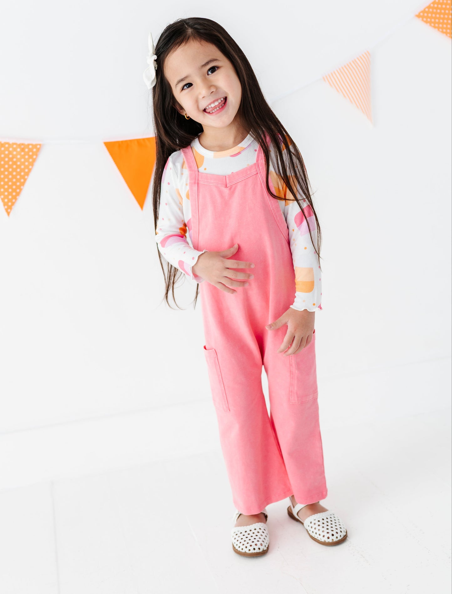 A young girl with long dark hair stands smiling in a pink overall-style romper over a white long-sleeve shirt with pastel accents, wearing white shoes. She is posed against a white studio background with orange and white bunting flags.