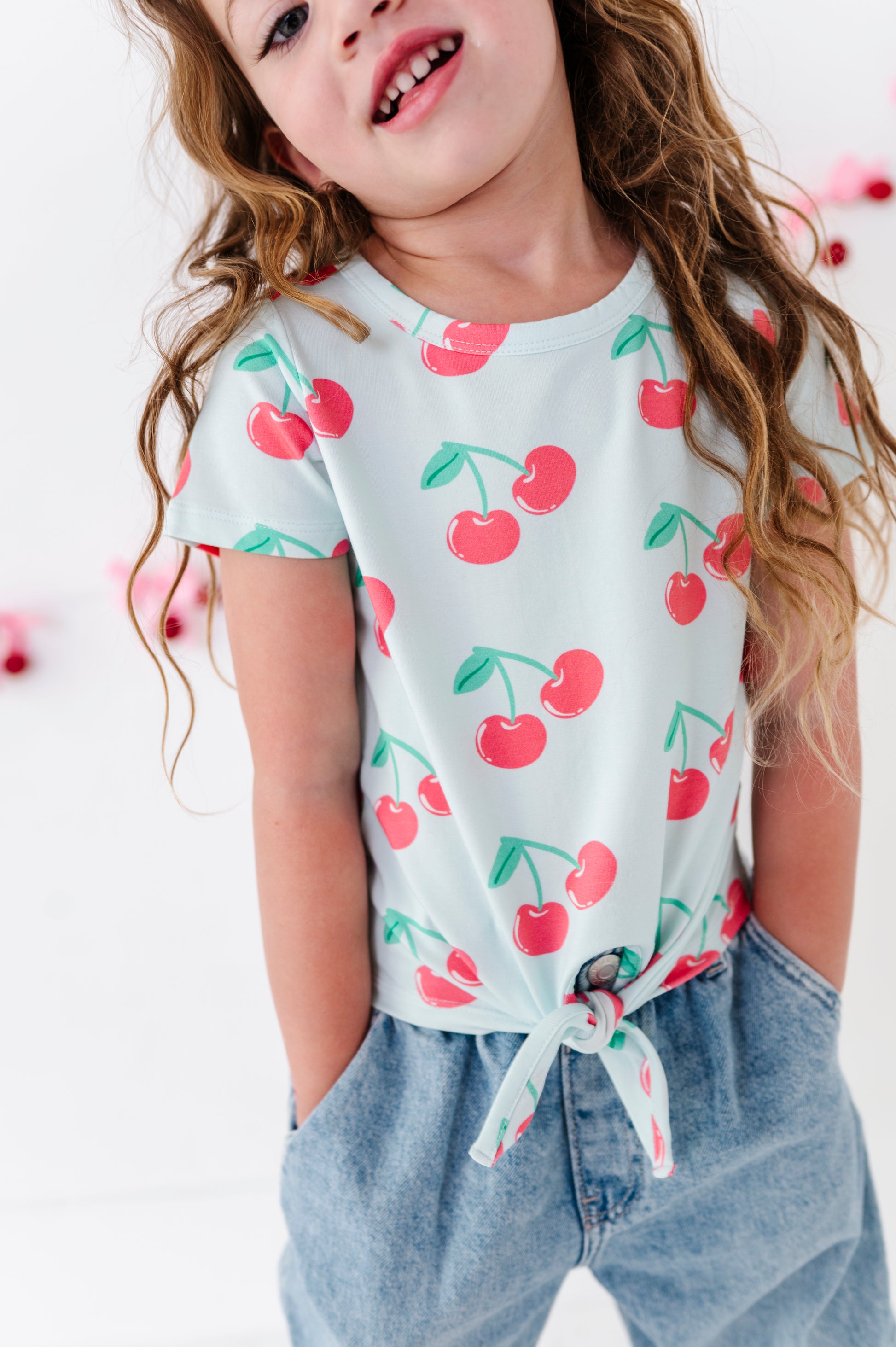 A young girl wearing a light blue short-sleeve t-shirt with a red cherry print and light blue jeans, shown from the chest up against a bright white background.