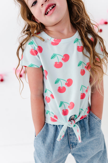 A young girl wearing a light blue short-sleeve t-shirt with a red cherry print and light blue jeans, shown from the chest up against a bright white background.