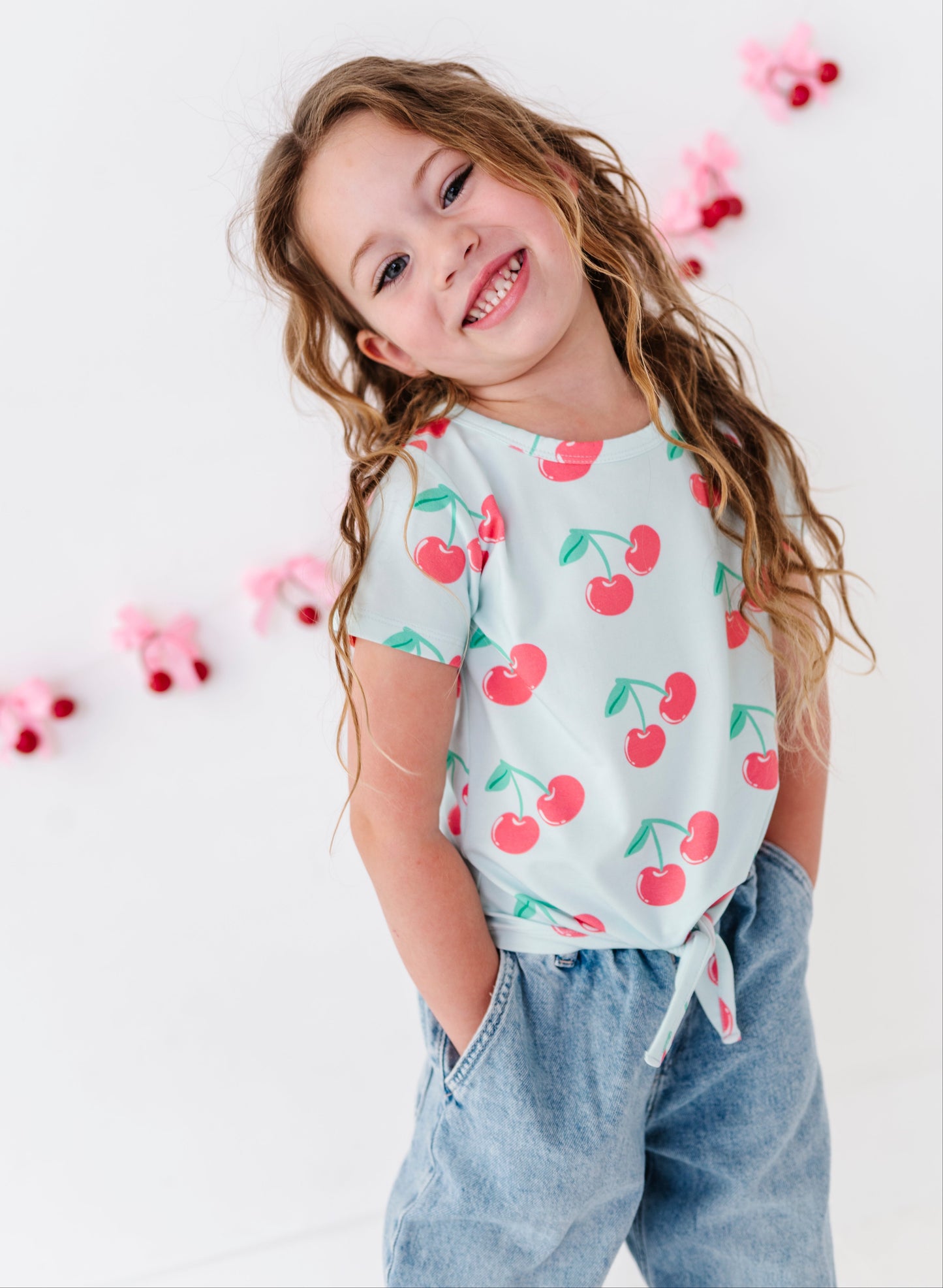 A young girl with long wavy light brown hair smiling and looking to the side, wearing a light blue short-sleeve t-shirt with a red cherry print and light blue jeans, standing against a white wall with pink cherry garlands.