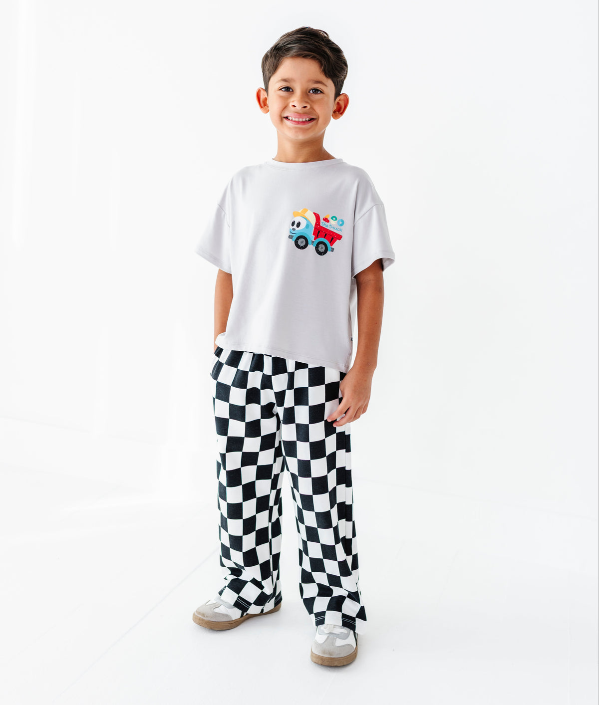 A young boy stands against a plain white studio background wearing a white t-shirt with a colorful truck graphic and black-and-white checkered pants.