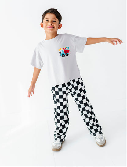 A young boy modeling a white t-shirt with a colorful cartoon truck graphic and black-and-white checkered pants, standing against a seamless white studio background.