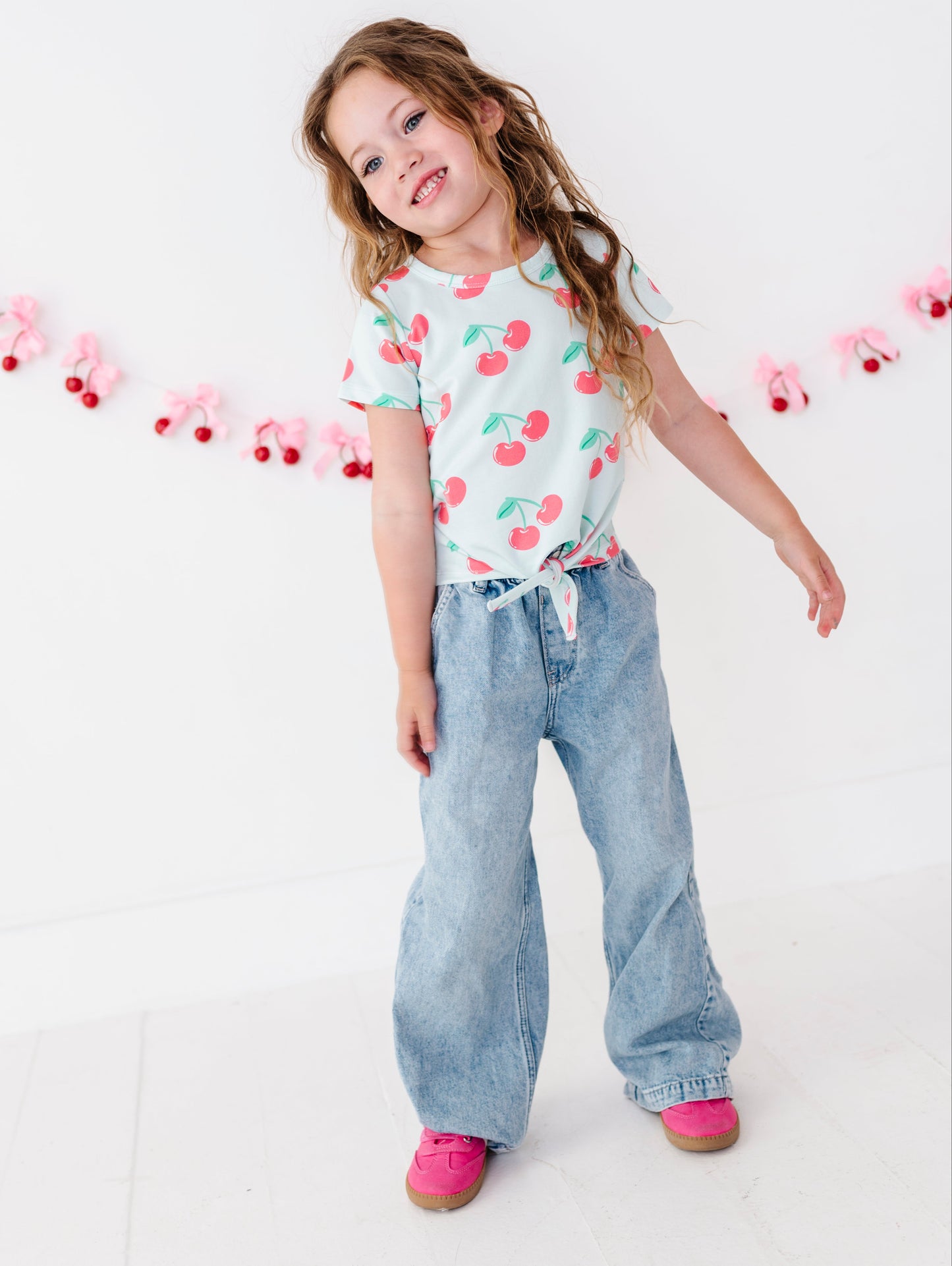 A young girl with long brown hair stands in a studio wearing a white t-shirt with a red and green cherry print, light blue wide-leg jeans, and pink shoes. She is posing against a white wall decorated with a pink and red bow garland.