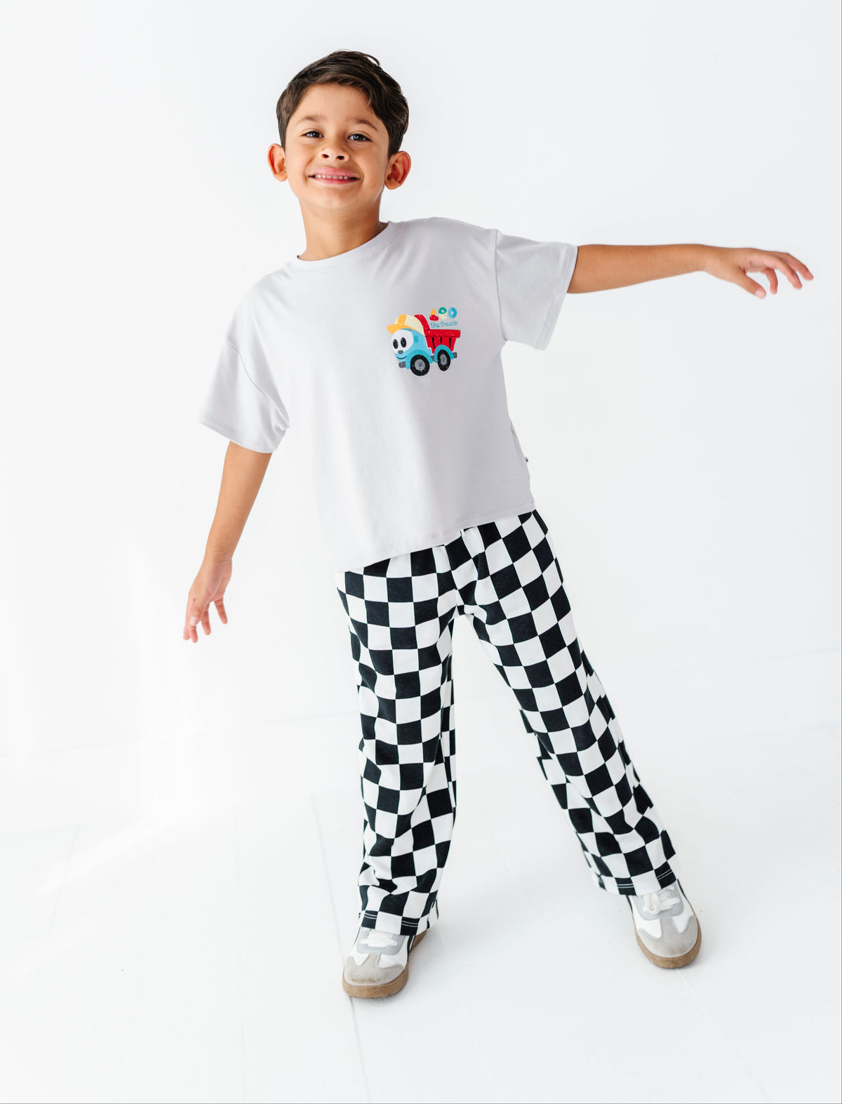 A young boy modeling a white t-shirt with a colorful cartoon truck graphic and black-and-white checkered pants, standing against a seamless white studio background.