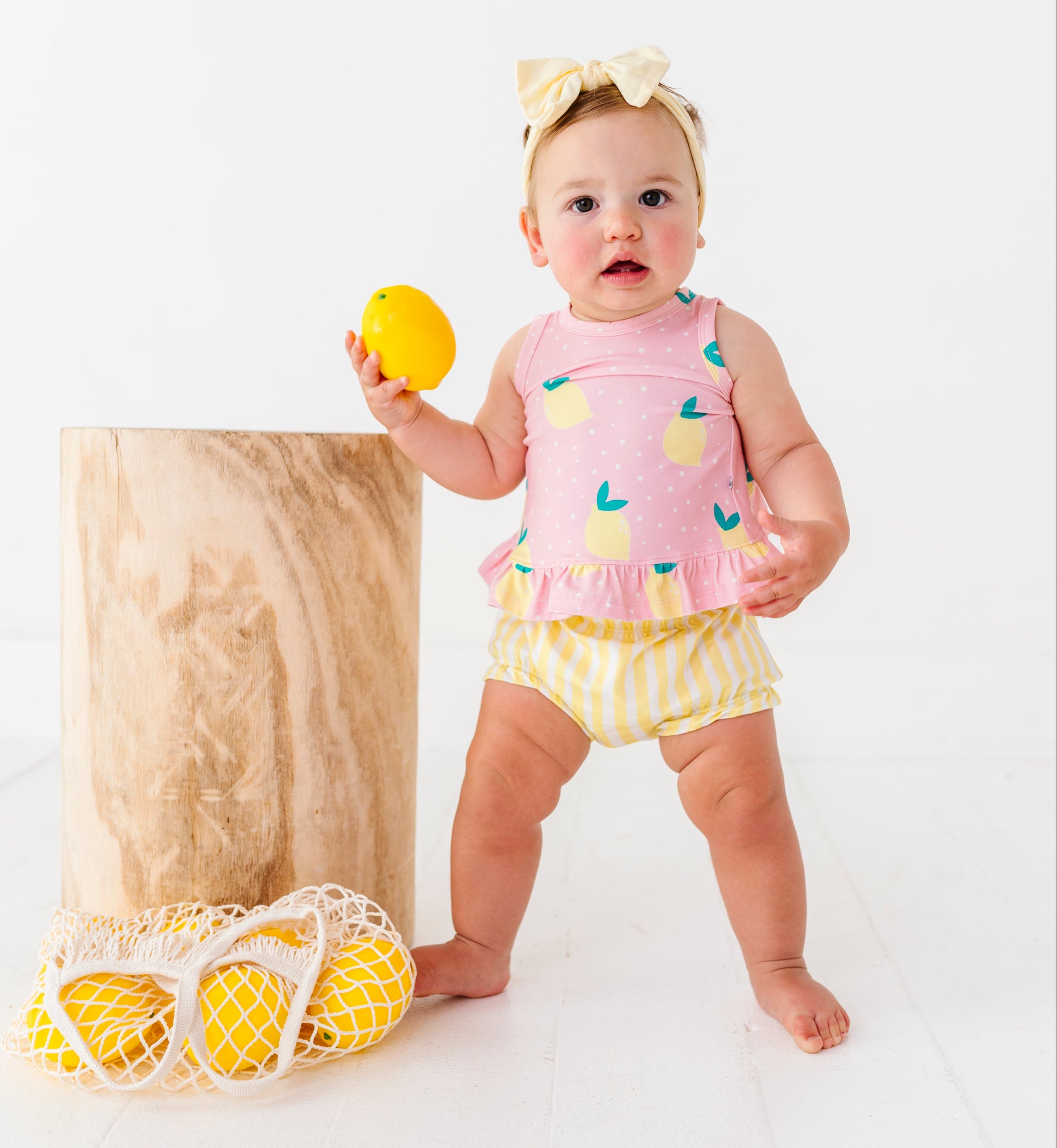Baby in a pink top and yellow bloomers holding a lemon toy on a white background