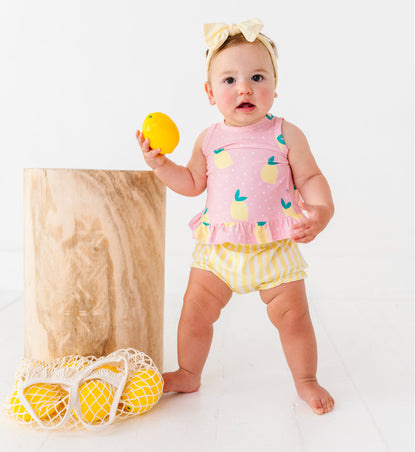 Baby in a pink top and yellow bloomers holding a lemon toy on a white background