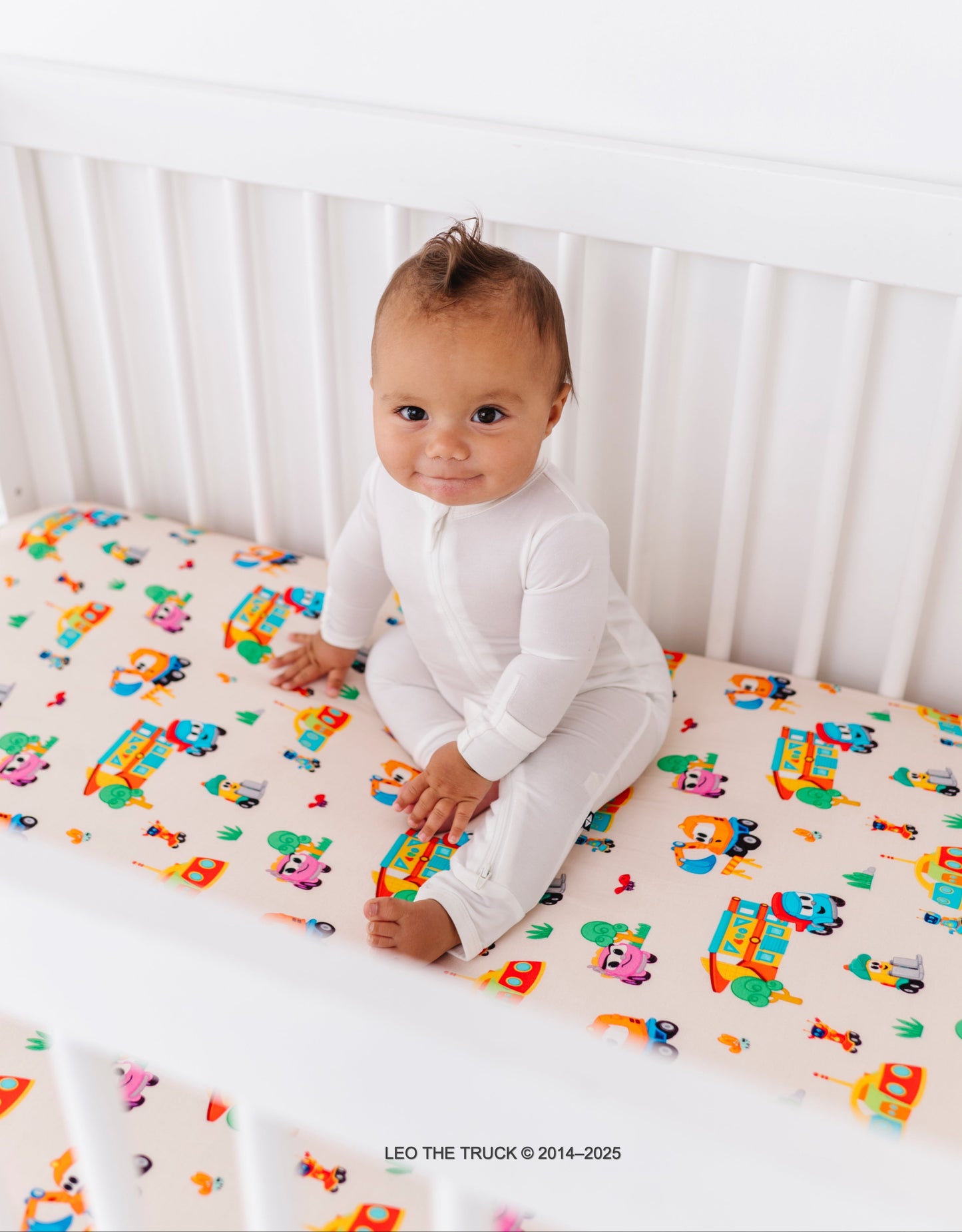 Baby sitting on a crib with colorful Leo the Truck bamboo fitted sheet 
