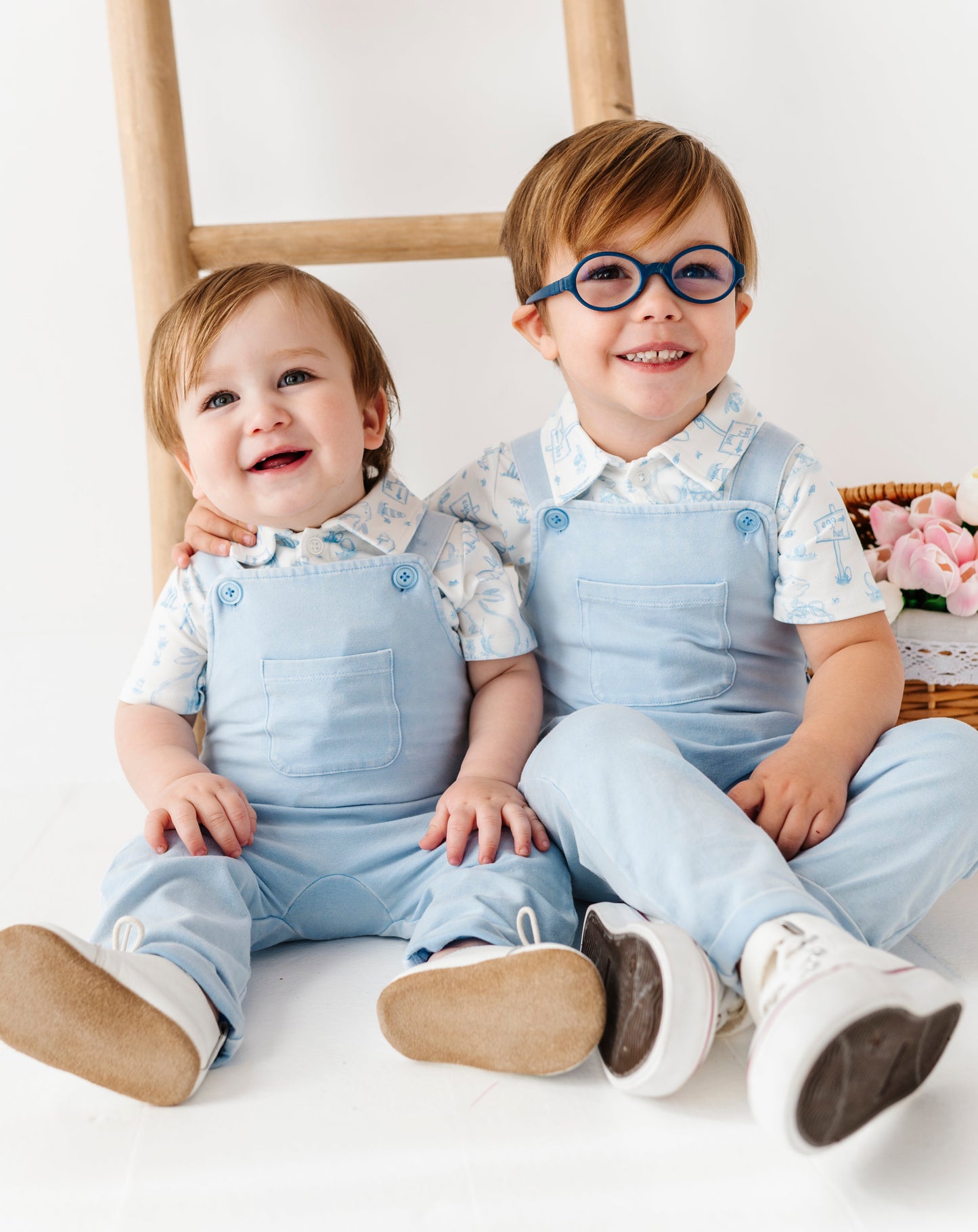 Two children in matching light blue bamboo overalls sitting on a white floor with a wooden ladder in the background.