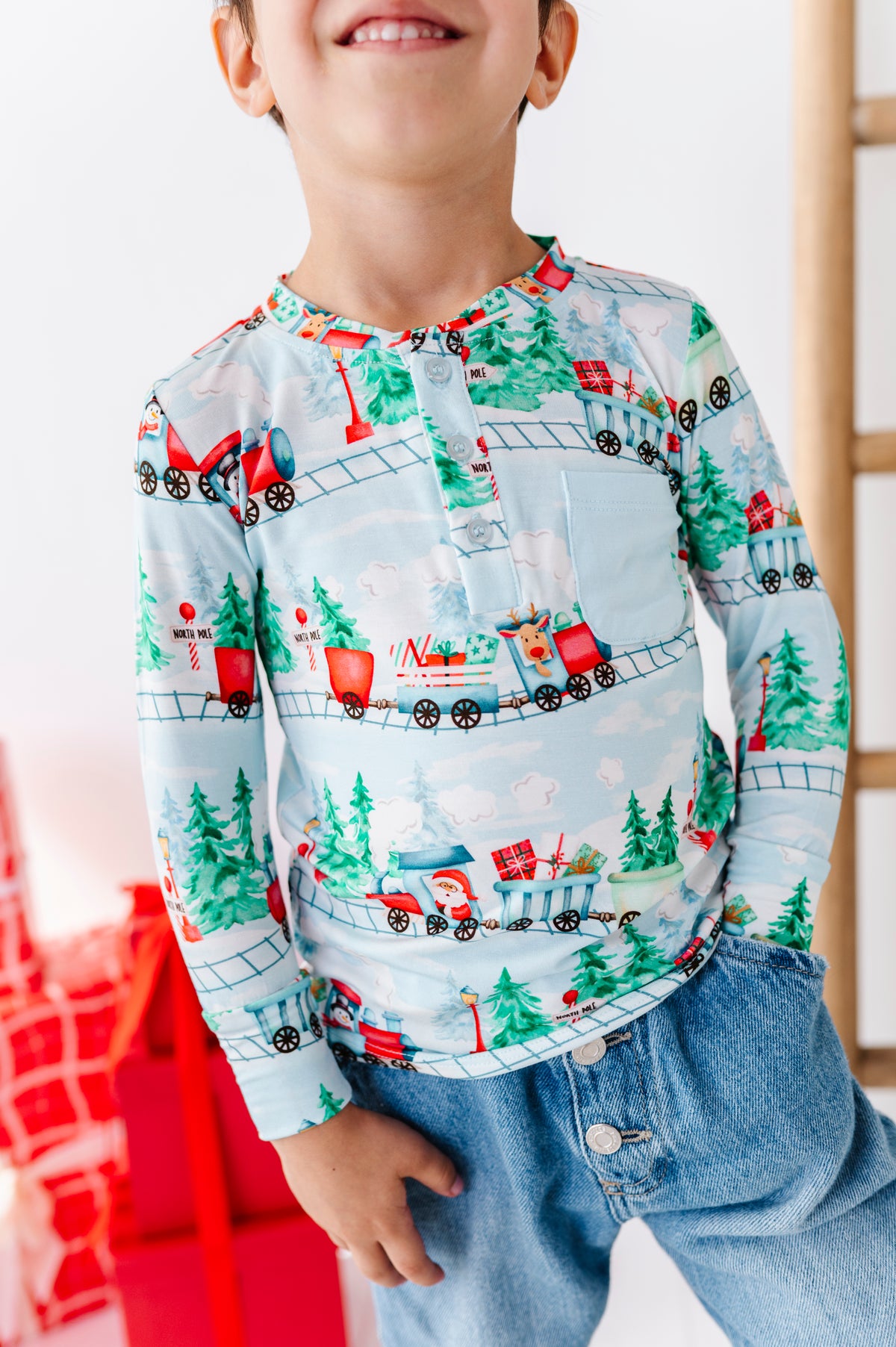 A child wearing a long-sleeve henley shirt with a festive Christmas train pattern, paired with blue jeans. The shirt features a light blue background with red, green, and white holiday illustrations of trains, pine trees, and snowflakes.