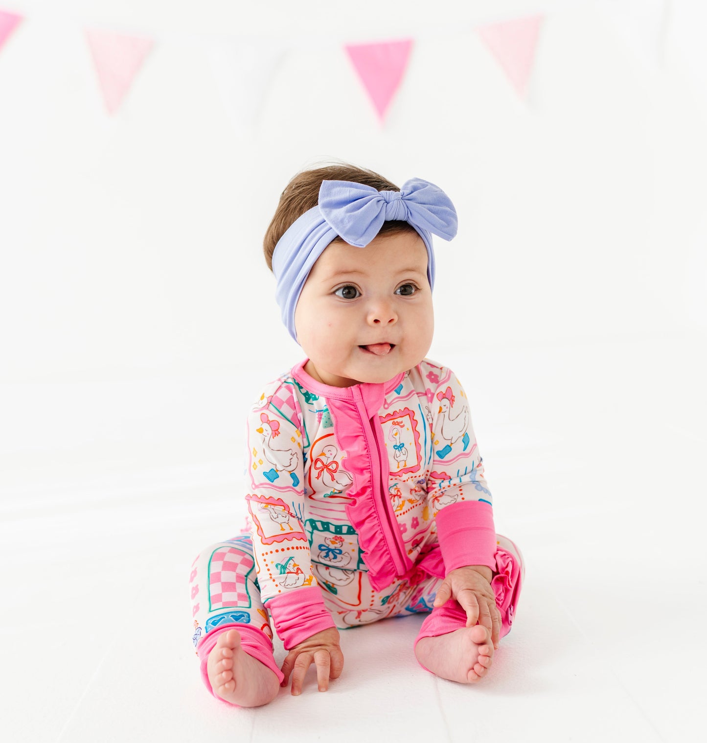 Baby wearing a colorful bamboo romper with pink accents and a headband, sitting on a white background with pink triangle flags.