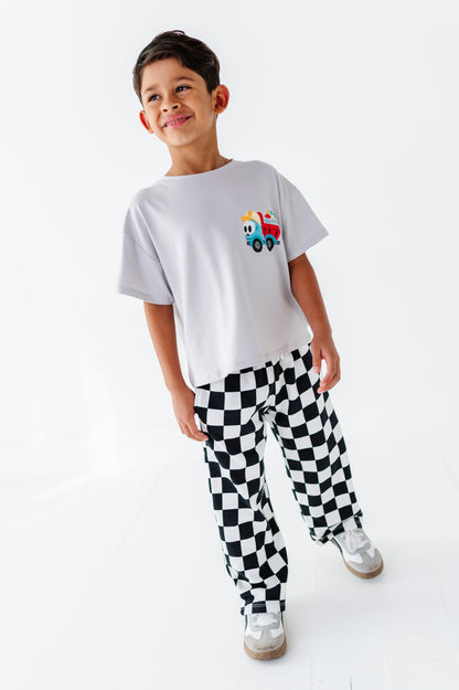 A young boy wearing a white t-shirt with a colorful fire truck graphic on the chest and black-and-white checkered pants, standing against a plain white studio background.