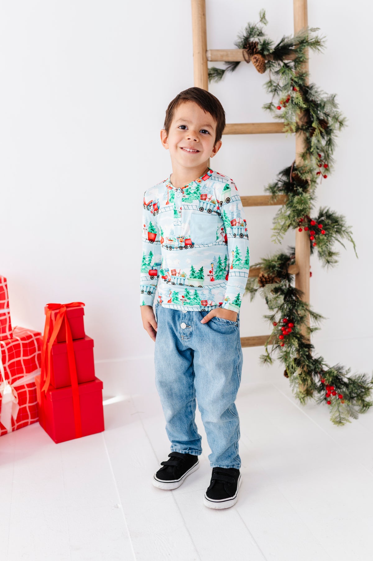 A child wearing a long-sleeve henley shirt with a festive Christmas train pattern, paired with blue jeans. The shirt features a light blue background with red, green, and white holiday illustrations of trains, pine trees, and snowflakes.