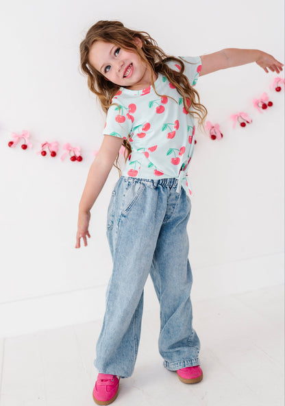 A young girl with long wavy brown hair poses in a white t-shirt with a red cherry print, paired with light blue wide-leg denim jeans and bright pink shoes, against a white wall with a pink and red cherry garland.