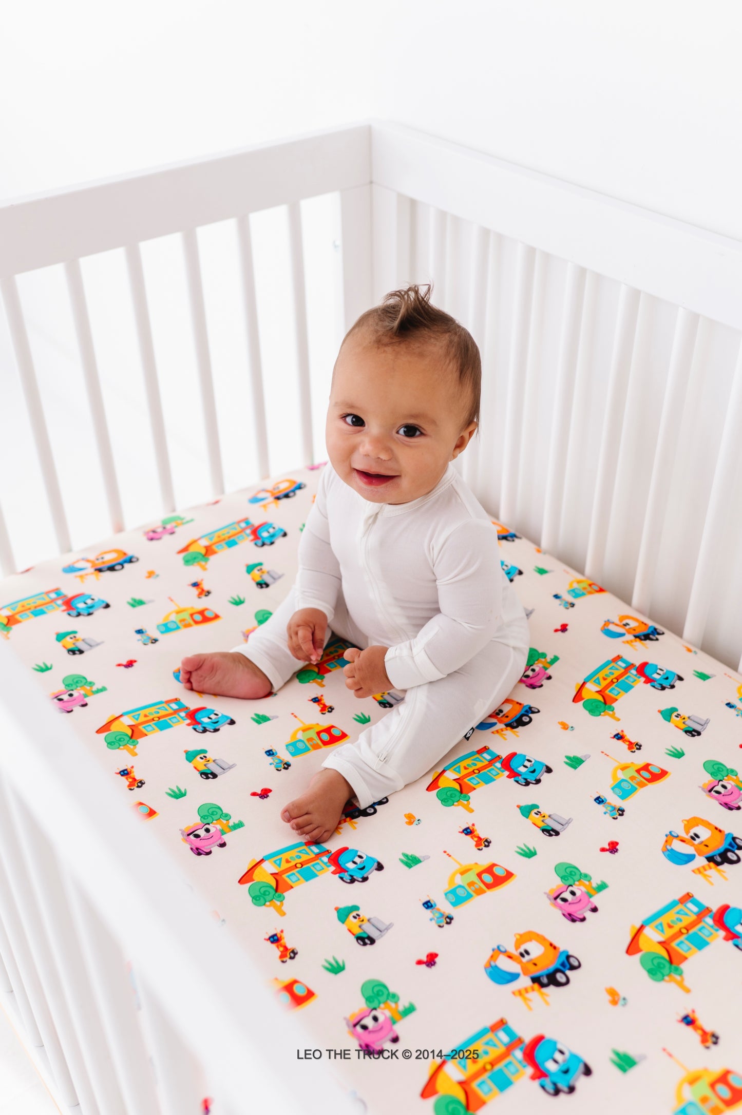 Baby sitting on a colorful Leo the Truck bamboo fitted sheet 