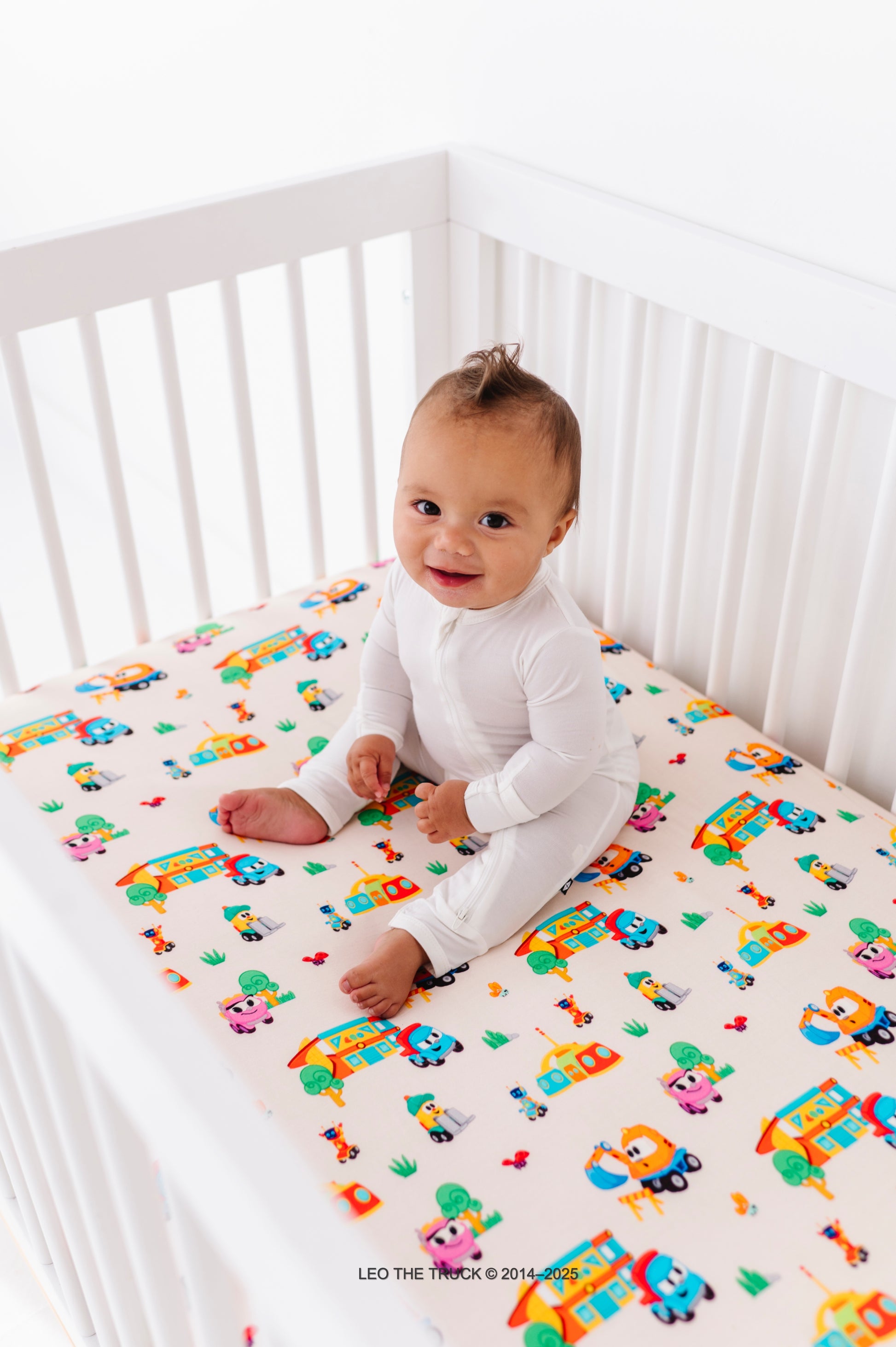 Baby sitting on a colorful Leo the Truck bamboo fitted sheet 