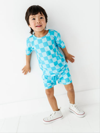 A young child wearing a light blue and white checkered short-sleeve t-shirt and matching shorts, paired with white canvas sneakers, standing against a clean white wall.