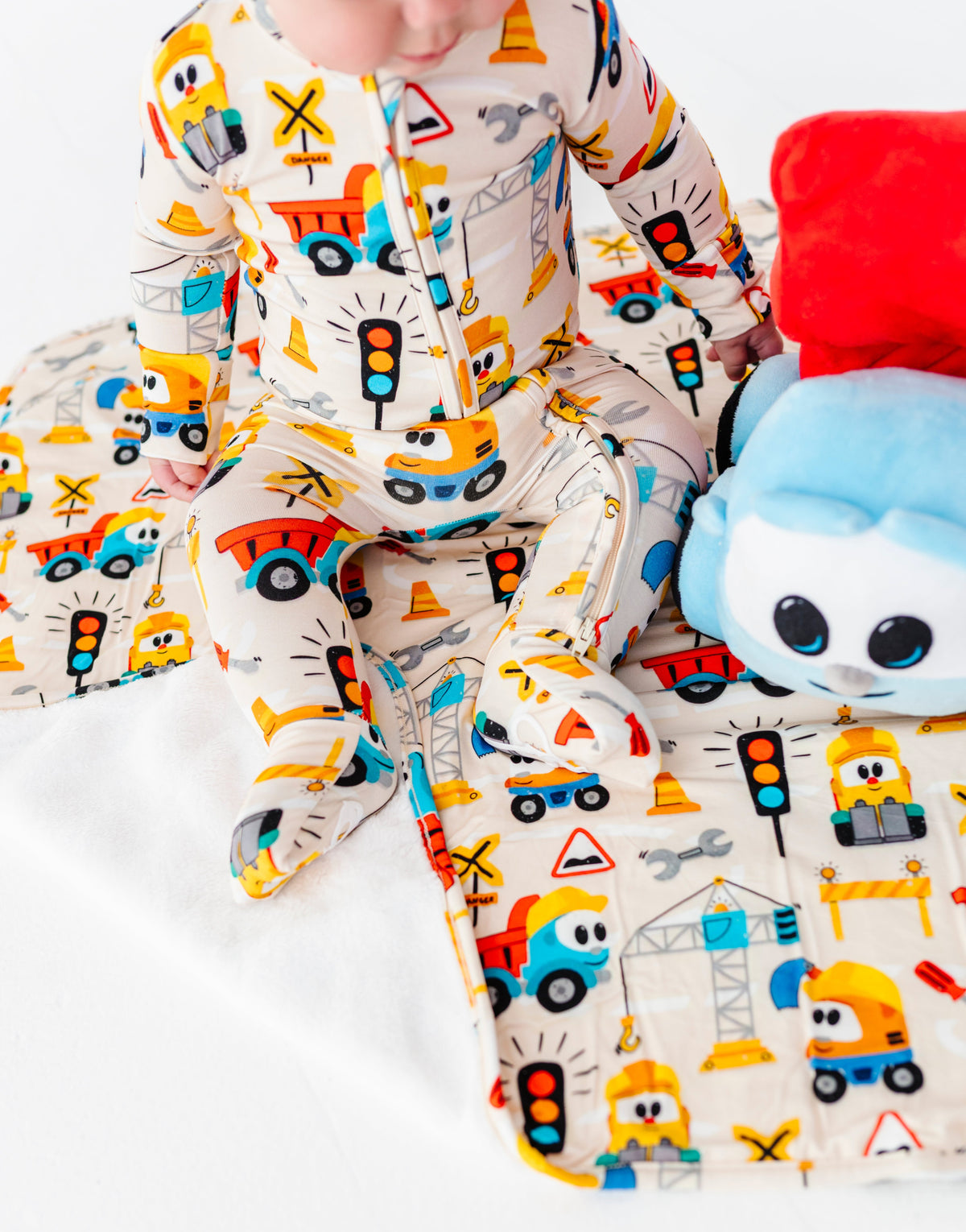 A baby wearing a cream-colored footed sleeper with a colorful construction vehicle print, sitting next to a plush toy on a white blanket.